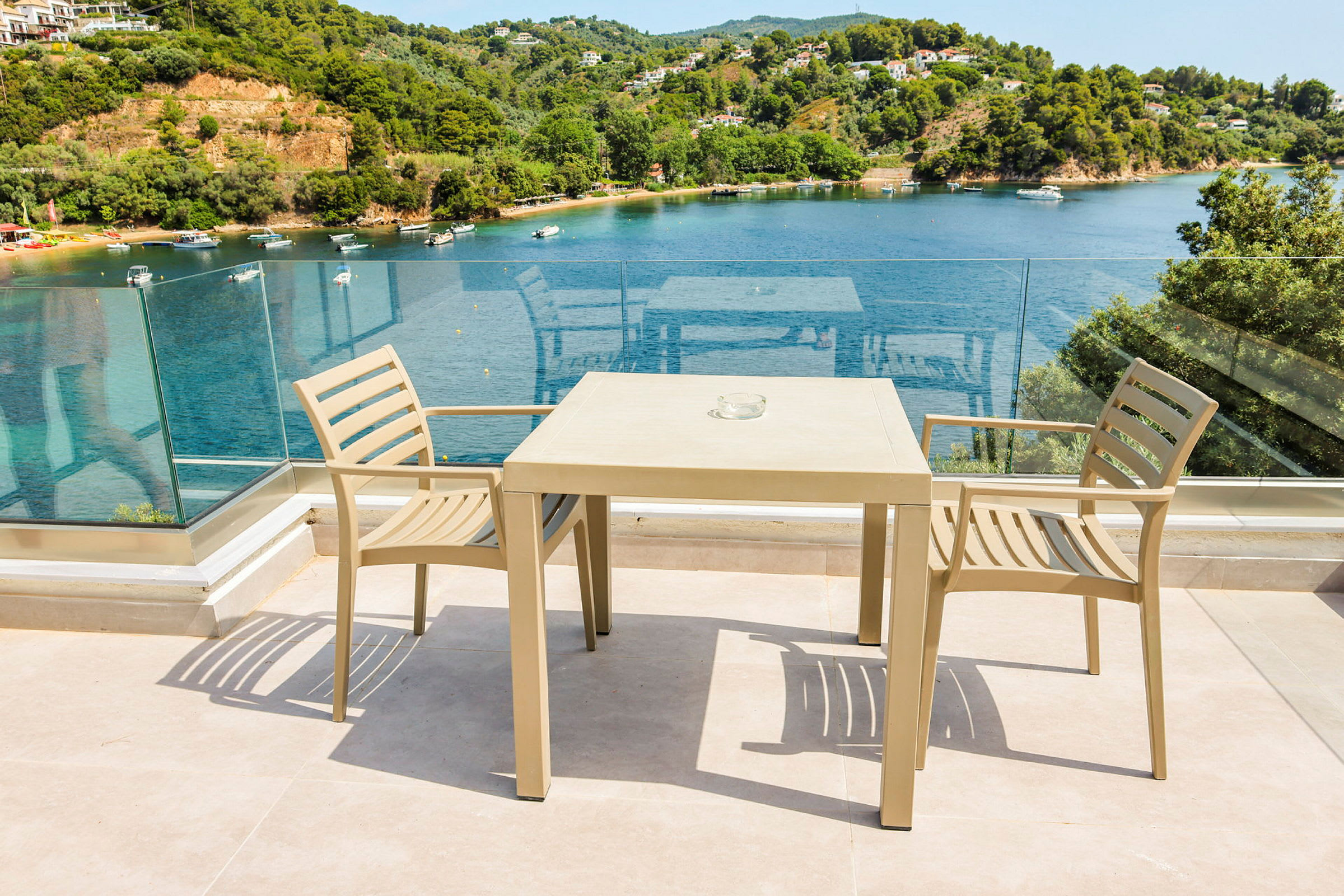 To beige stoler og et bord på en terrasse med glassrekkverk og utsikt over en bukt med båter og grøntområder. (AI-generert tekst)