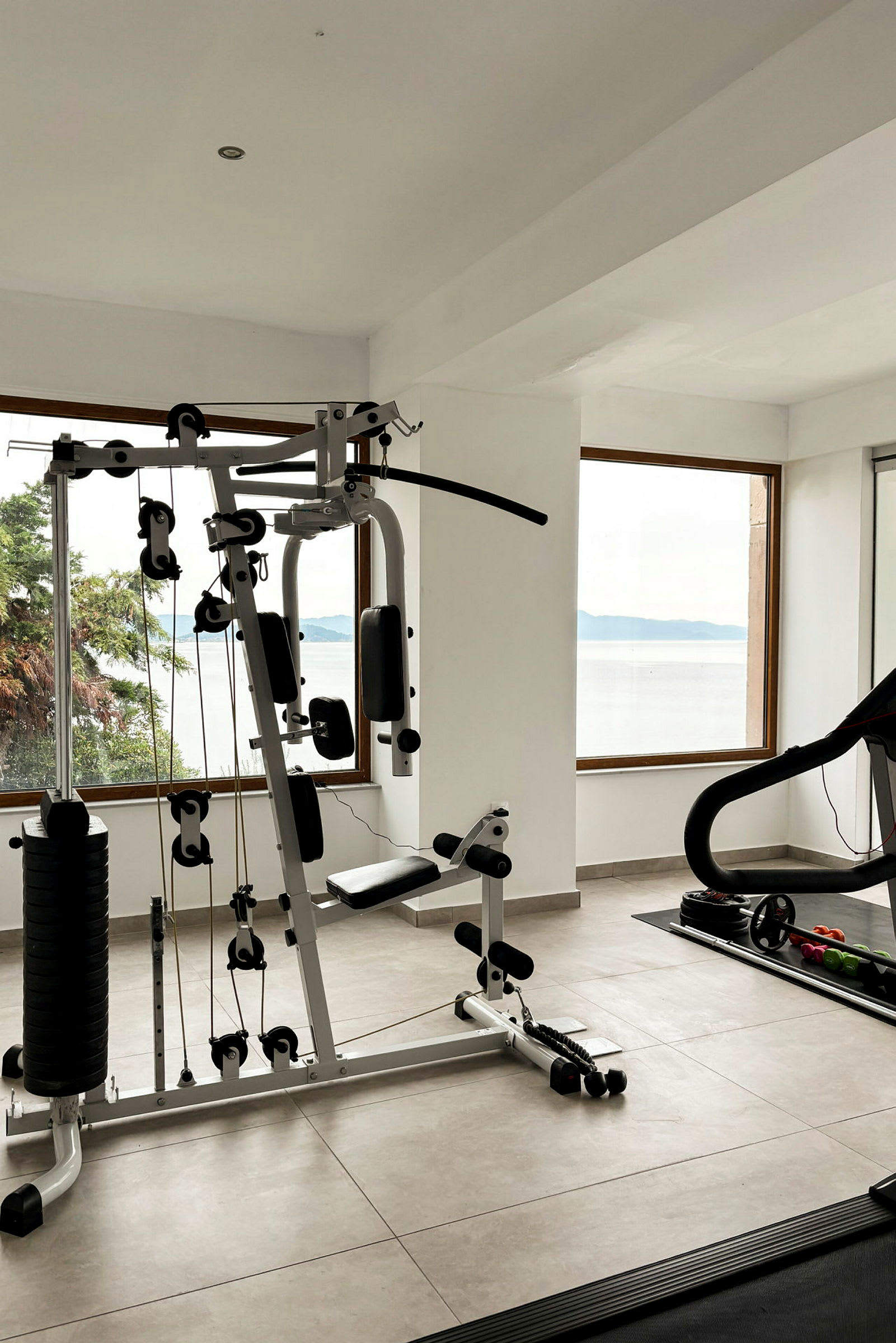 Et treningsstudio med treningsutstyr, inkludert en multigym og et tredemølle, med vinduer som vender mot sjøutsikten. (AI-generert tekst)