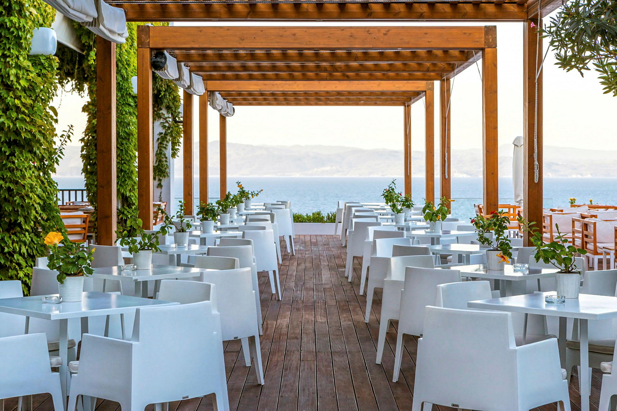 Utendørs restaurant med hvite bord og stoler under trepergola, omgitt av grønt. (AI-generert tekst)