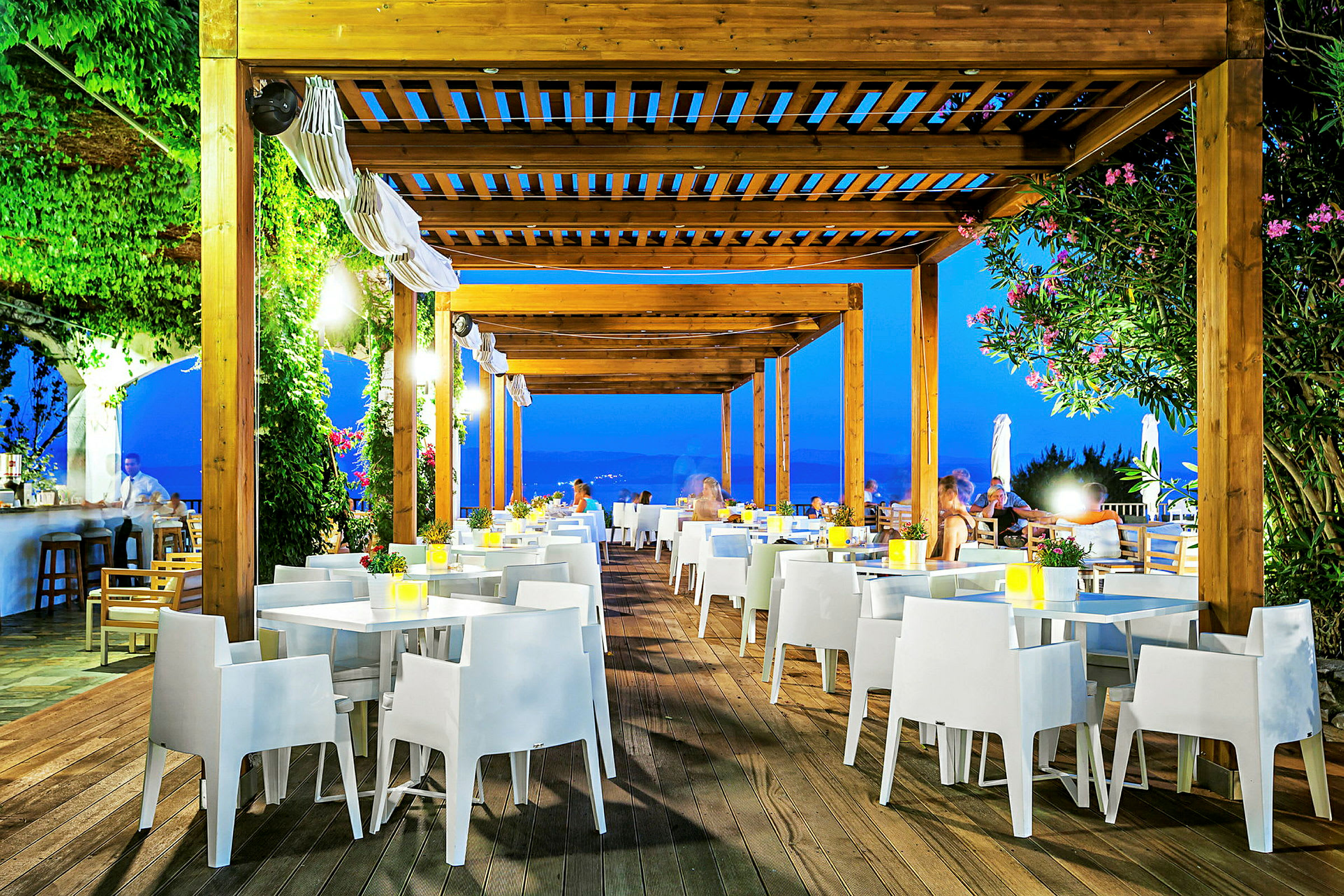 Utendørsrestaurant med trepergola, hvite stoler og bord, omgitt av grøntområder og kveldsbelysning. (AI-generert tekst)