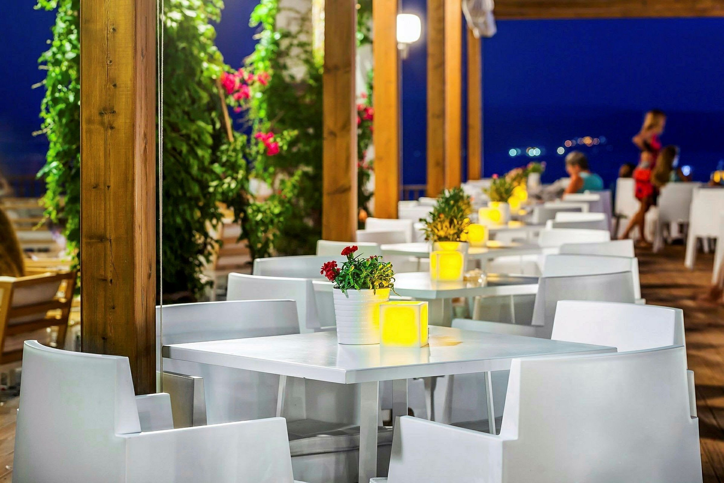 En utendørsrestaurant med hvite bord og stoler, dekorert med blomster og lys på kvelden. (AI-generert tekst)