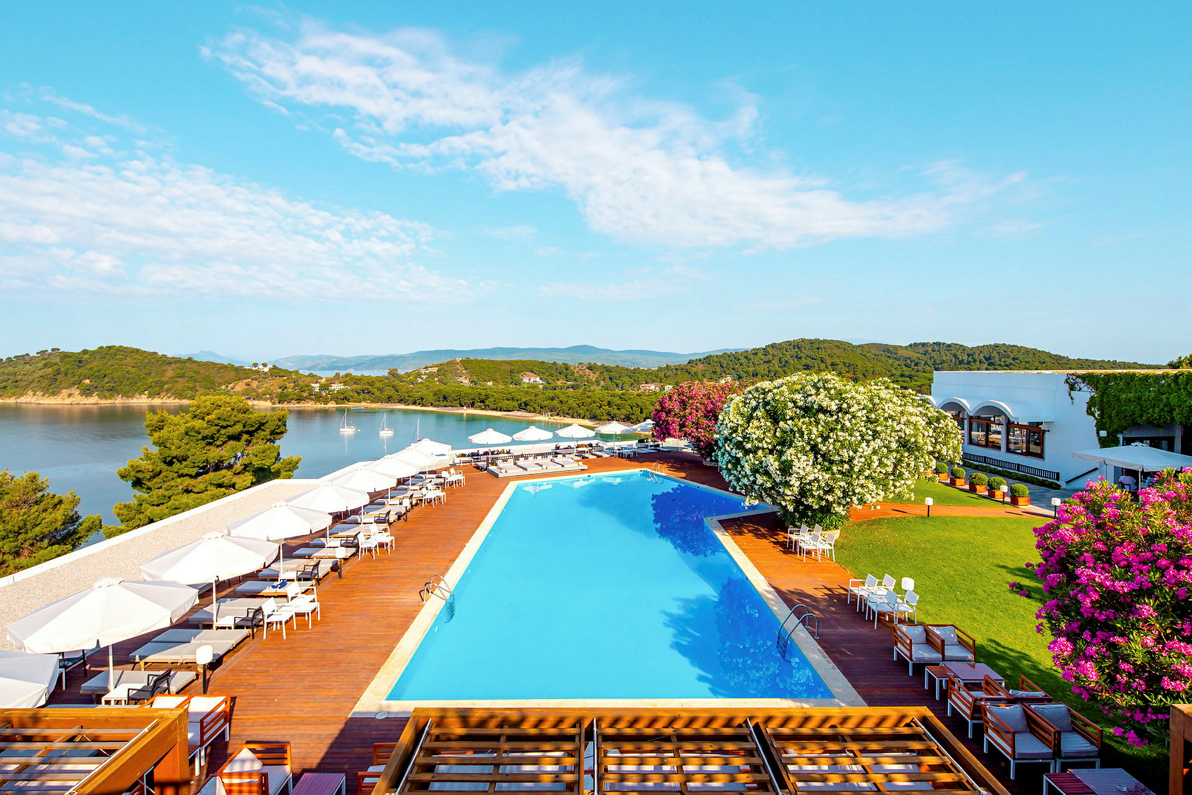 Utendørsområde med basseng og terrasse ved sjøen, omgitt av frodig vegetasjon og bygninger i bakgrunnen. (AI-generert tekst)