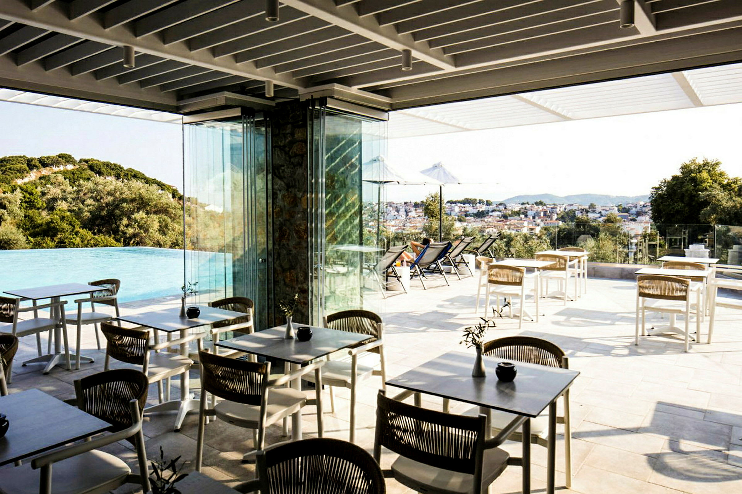 En utendørs terrasse med bord og stoler nær et infinitybasseng, med utsikt over et landskap og en by. (AI-generert tekst)