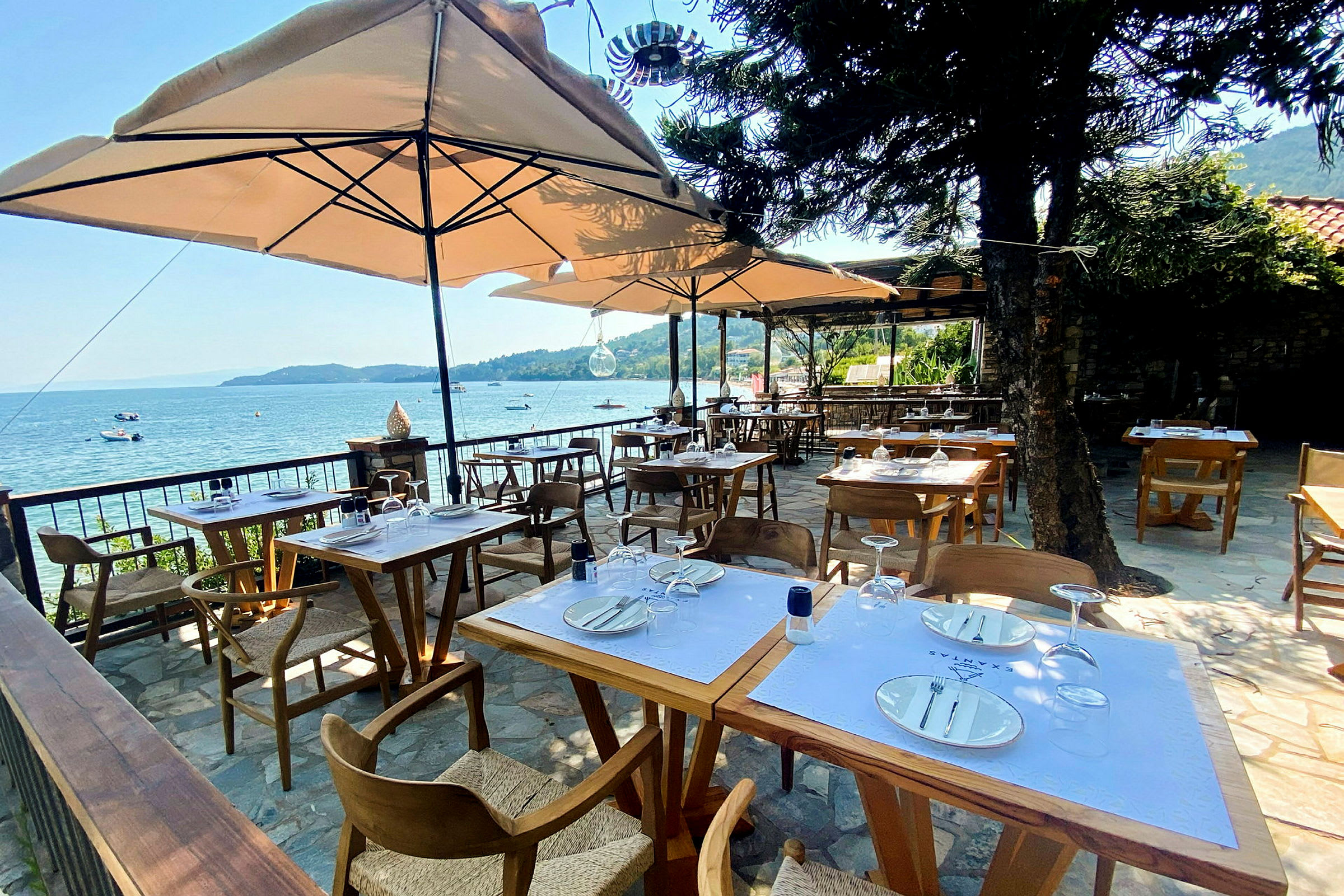 Bilde av en utendørs restaurant med bord og stoler under parasoller, med utsikt over havet i bakgrunnen. (AI-generert tekst)