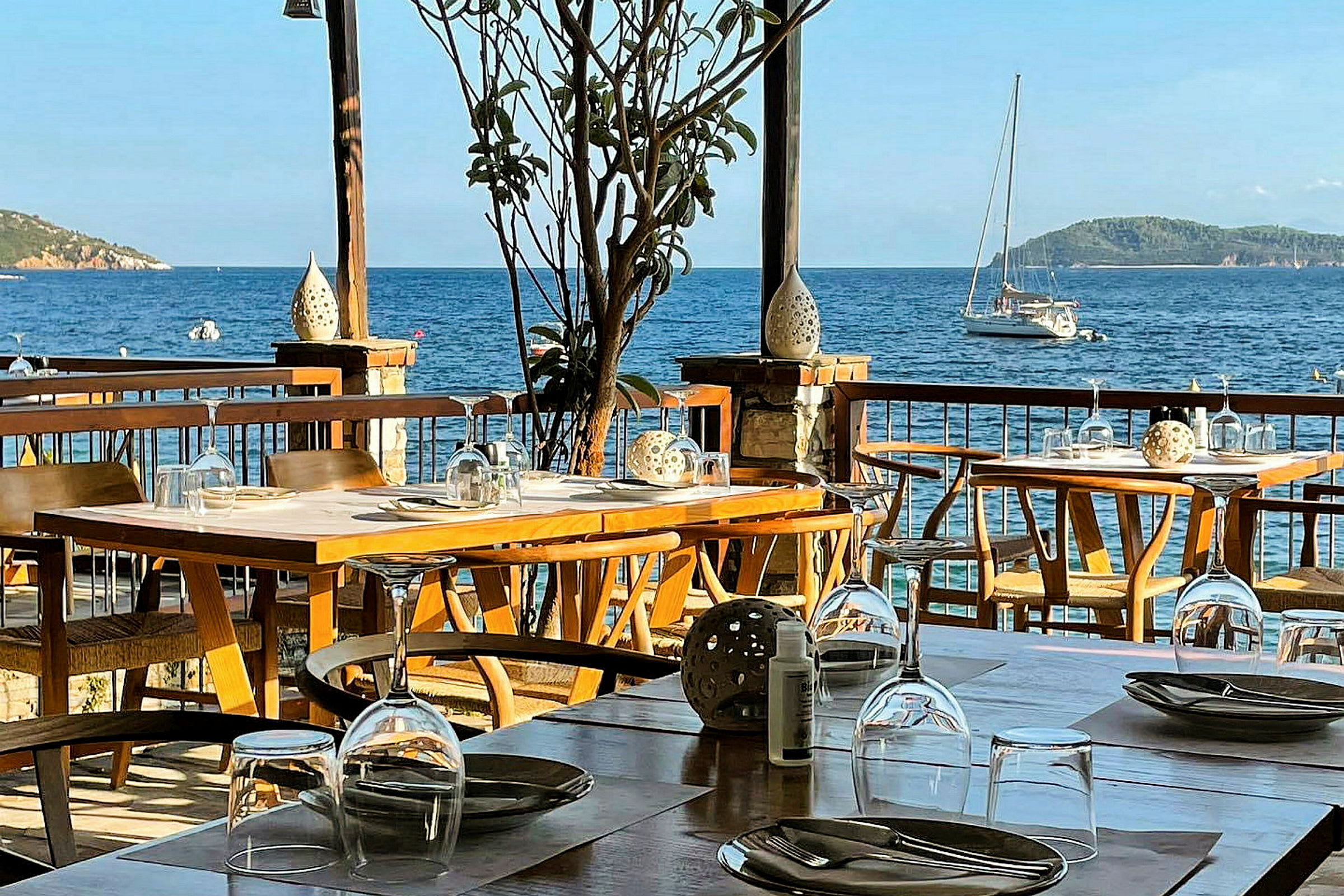 Uteområde med dekkede bord og utsikt over havet med en seilbåt. (AI-generert tekst)