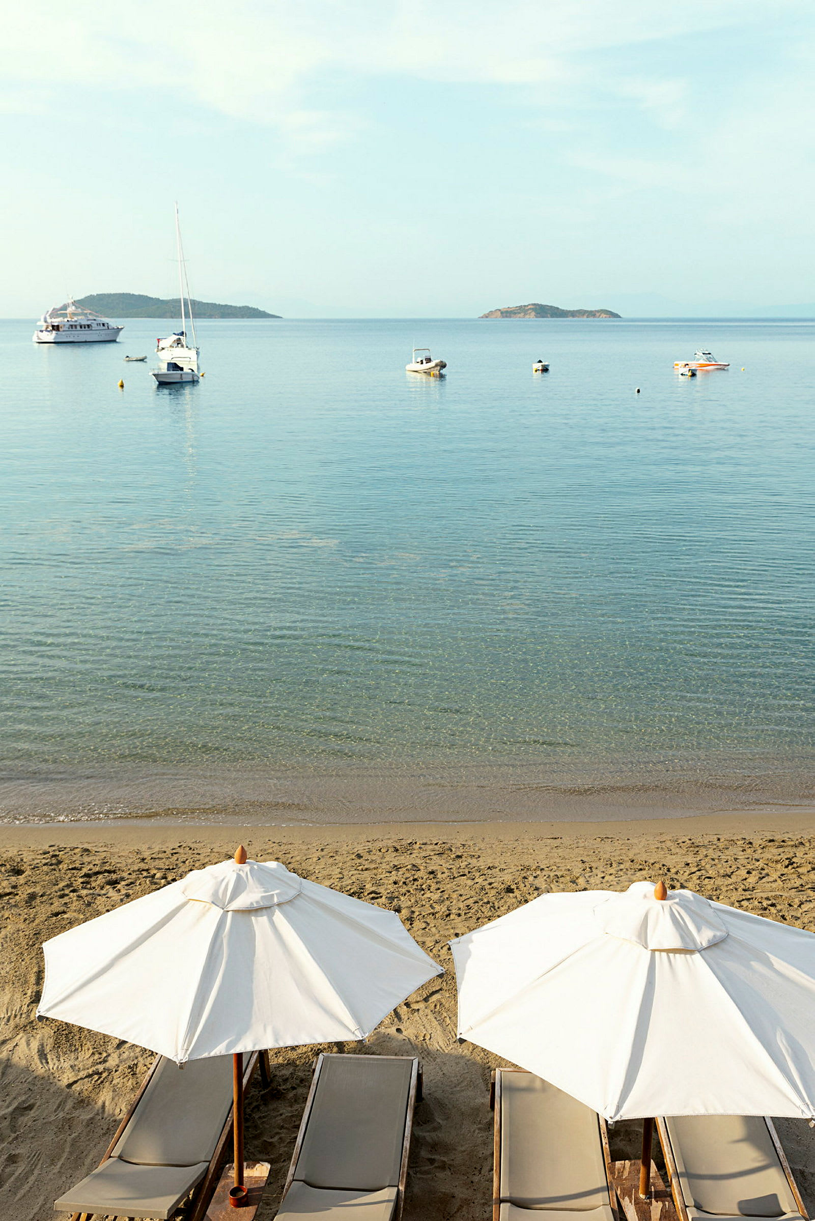 Strand med solsenger og parasoller, hav med båter og øyer i bakgrunnen. (AI-generert tekst)