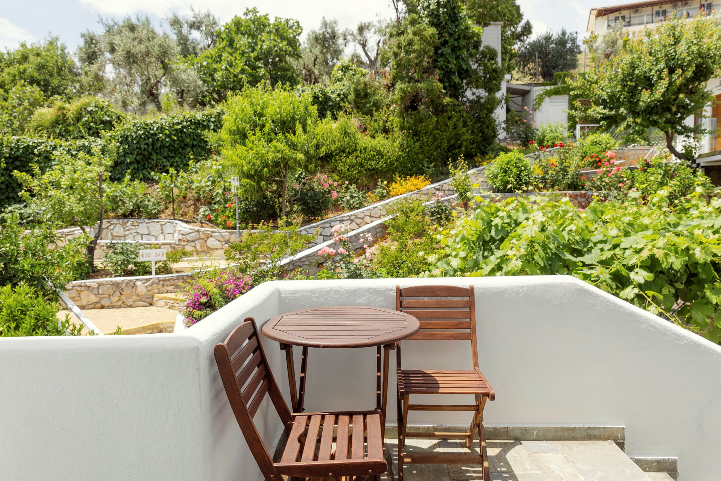 En liten terrasse med to trestoler og et rundt bord, omgitt av grøntområder og en steinbelagt hage. (AI-generert tekst)