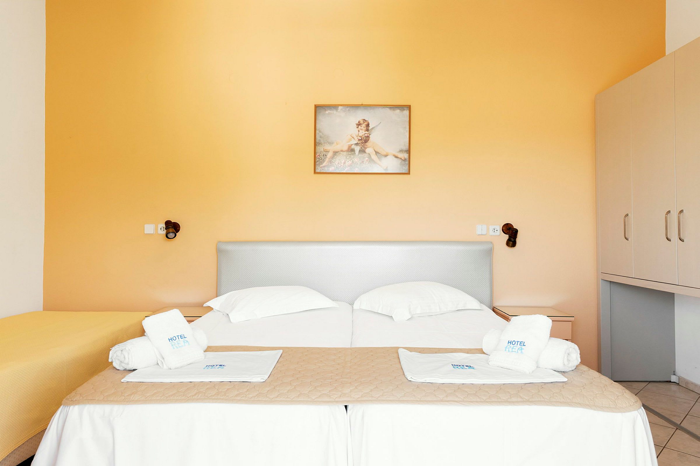Et hotellrom med to senger kledd i hvitt sengetøy og håndklær plassert oppå. Veggene er malt i oransje farge. (AI-generert tekst)