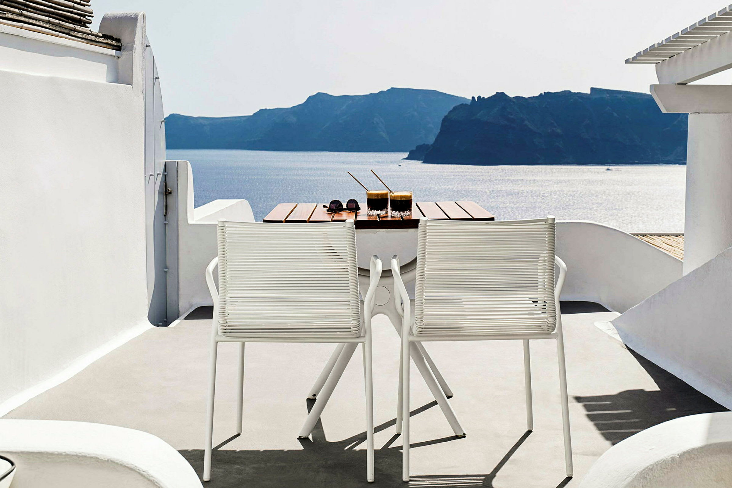 Et bord med to drikker og et par solbriller står på en terrasse med to hvite stoler. Utsikten viser havet og landskap i bakgrunnen. (AI-generert tekst)