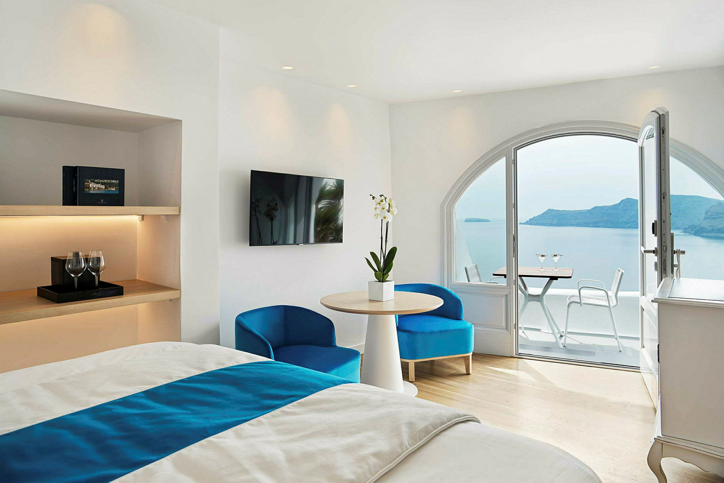 Hotellrom med lyse hvite vegger, møblert med en seng, bord og stoler, og en balkong med utsikt over havet og en øy. (AI-generert tekst)