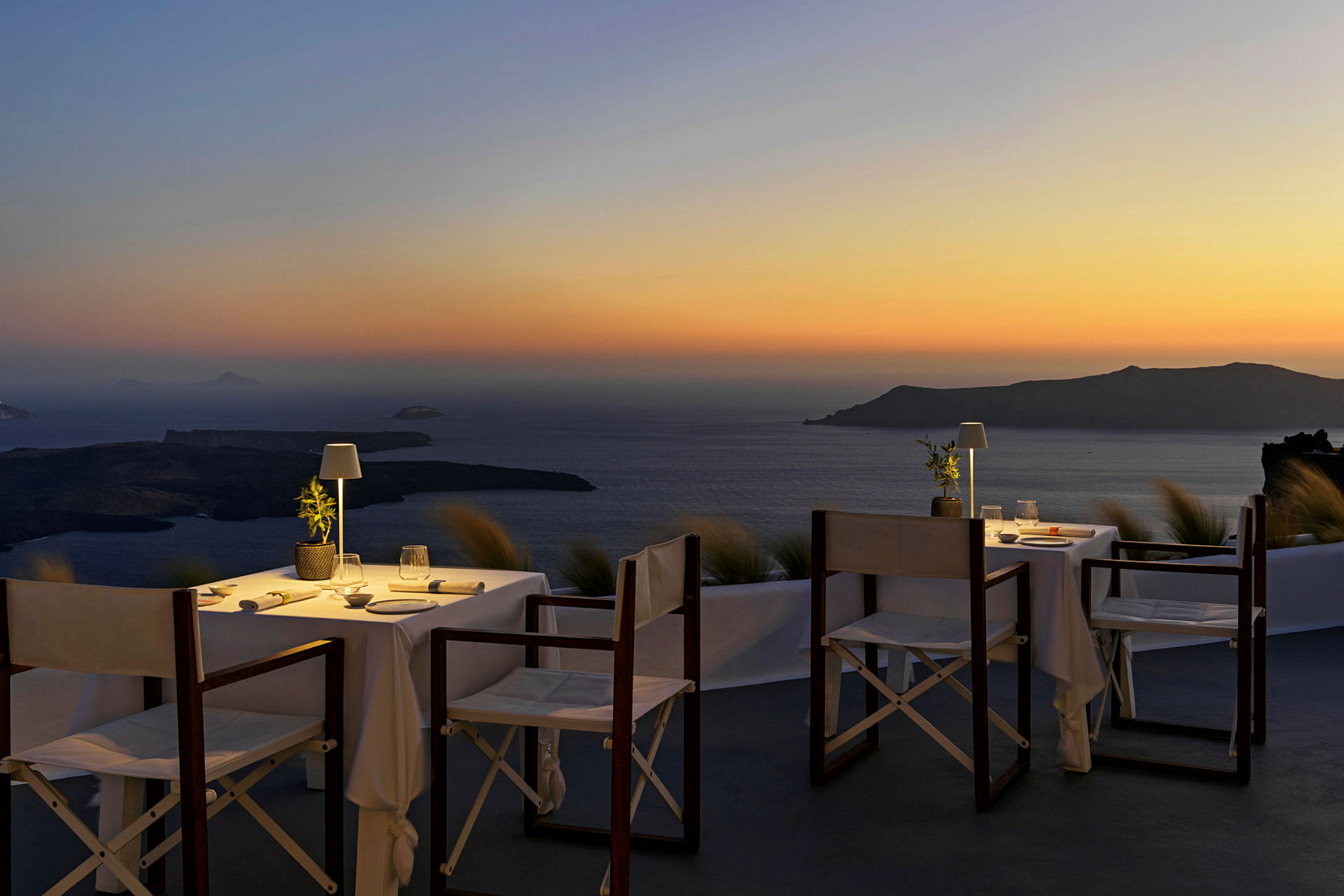 To dekkede bord med stoler på en terrasse, med utsikt over havet og en øy ved solnedgang. (AI-generert tekst)