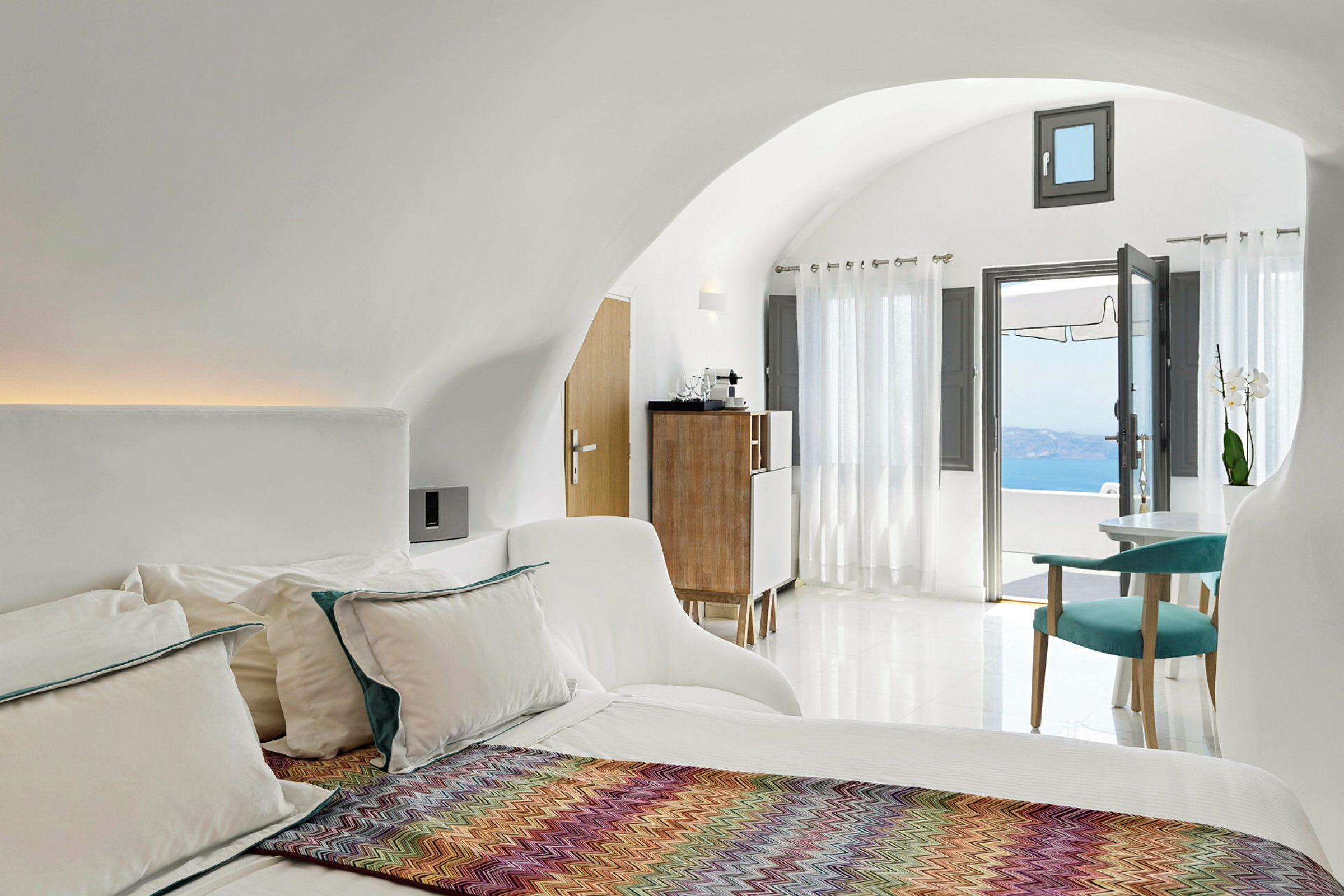 Et lyst og moderne hotellrom med en seng, fargerikt pledd, sittegruppe og store vinduer med utsikt over havet. (AI-generert tekst)