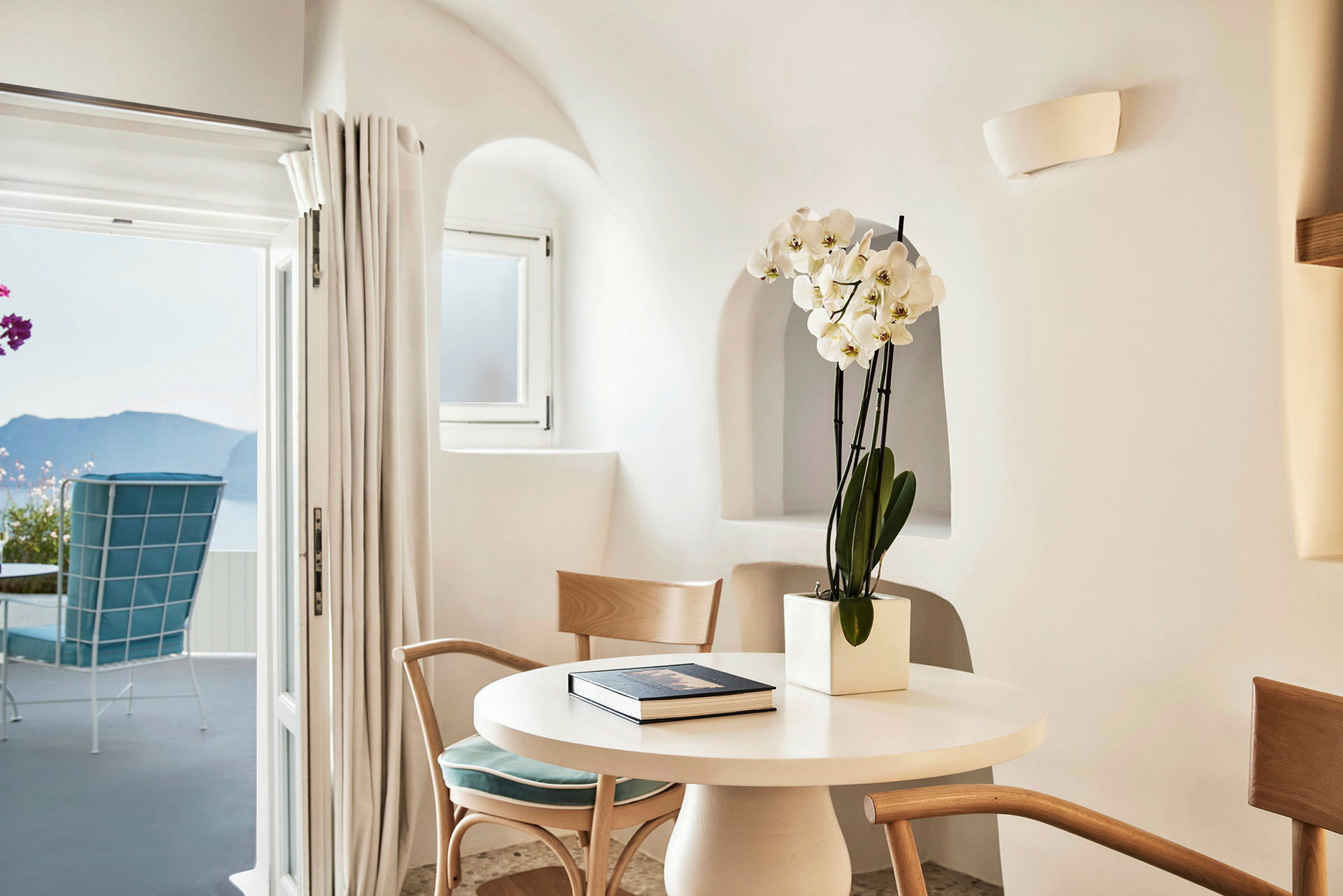 Rom i lys og moderne stil med runde vinduer, blomster i potte på bordet og en utsikt over havet gjennom en åpen dør. (AI-generert tekst)