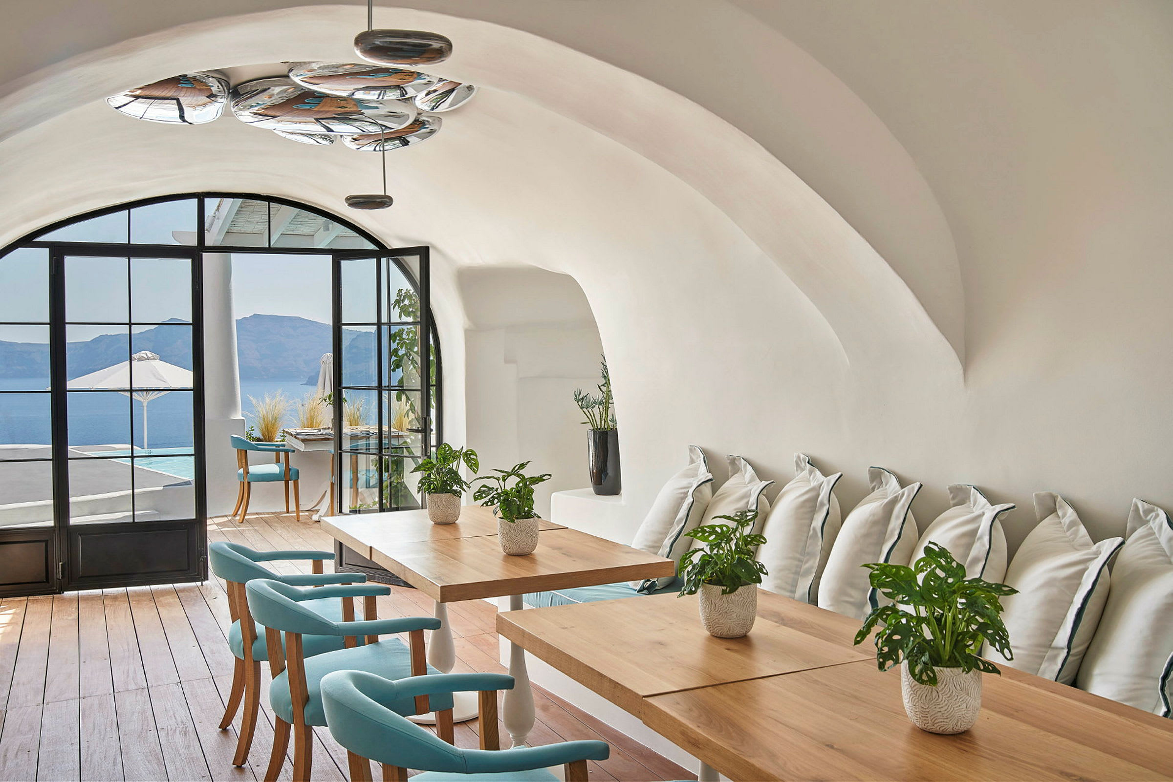 Et elegant interiør med hvelvede tak, trebord og turkise stoler. Store vinduer gir utsikt over en terrasse og havet. (AI-generert tekst)