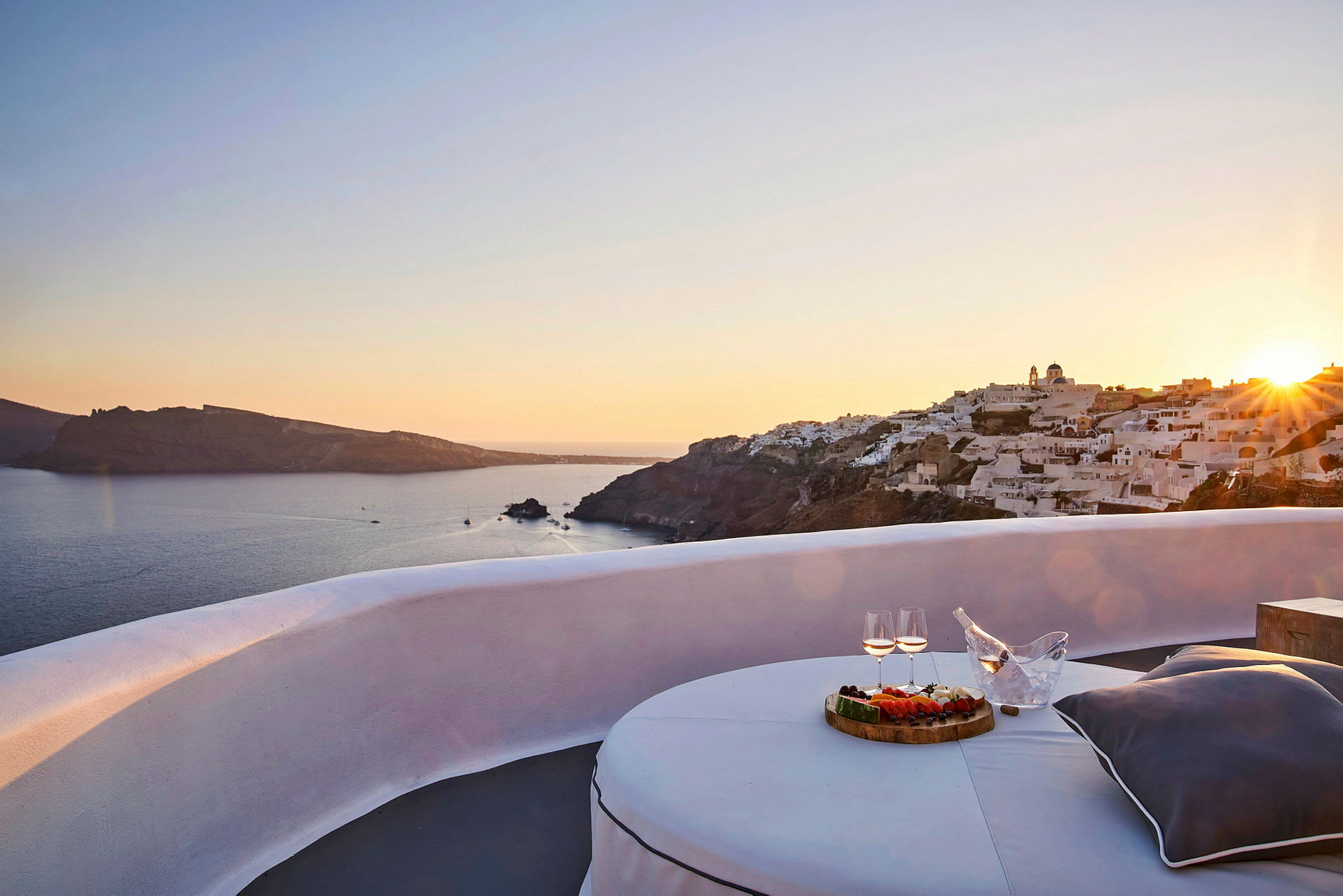 Utsikt over en solnedgang ved sjøen med hvite bygninger på en klippe og en terrasse med bord, mat og drikke i forgrunnen. (AI-generert tekst)