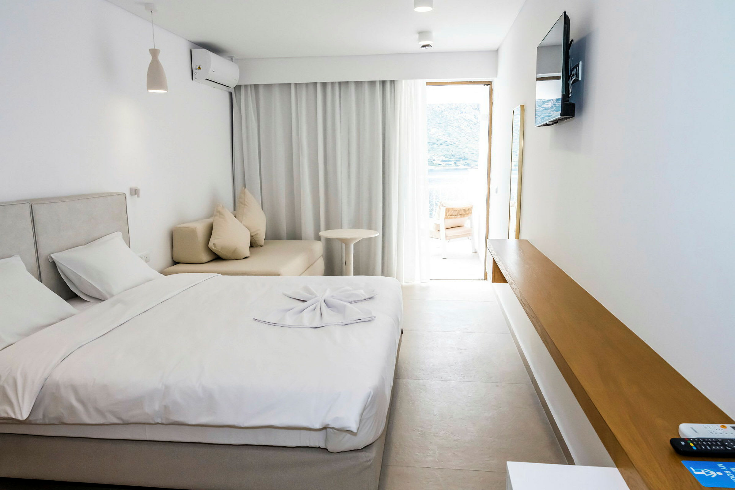 Et hotellrom med en seng, sofa, et bord og en TV på veggen. Et vindu med lyse gardiner fører til en terrasse. (AI-generert tekst)