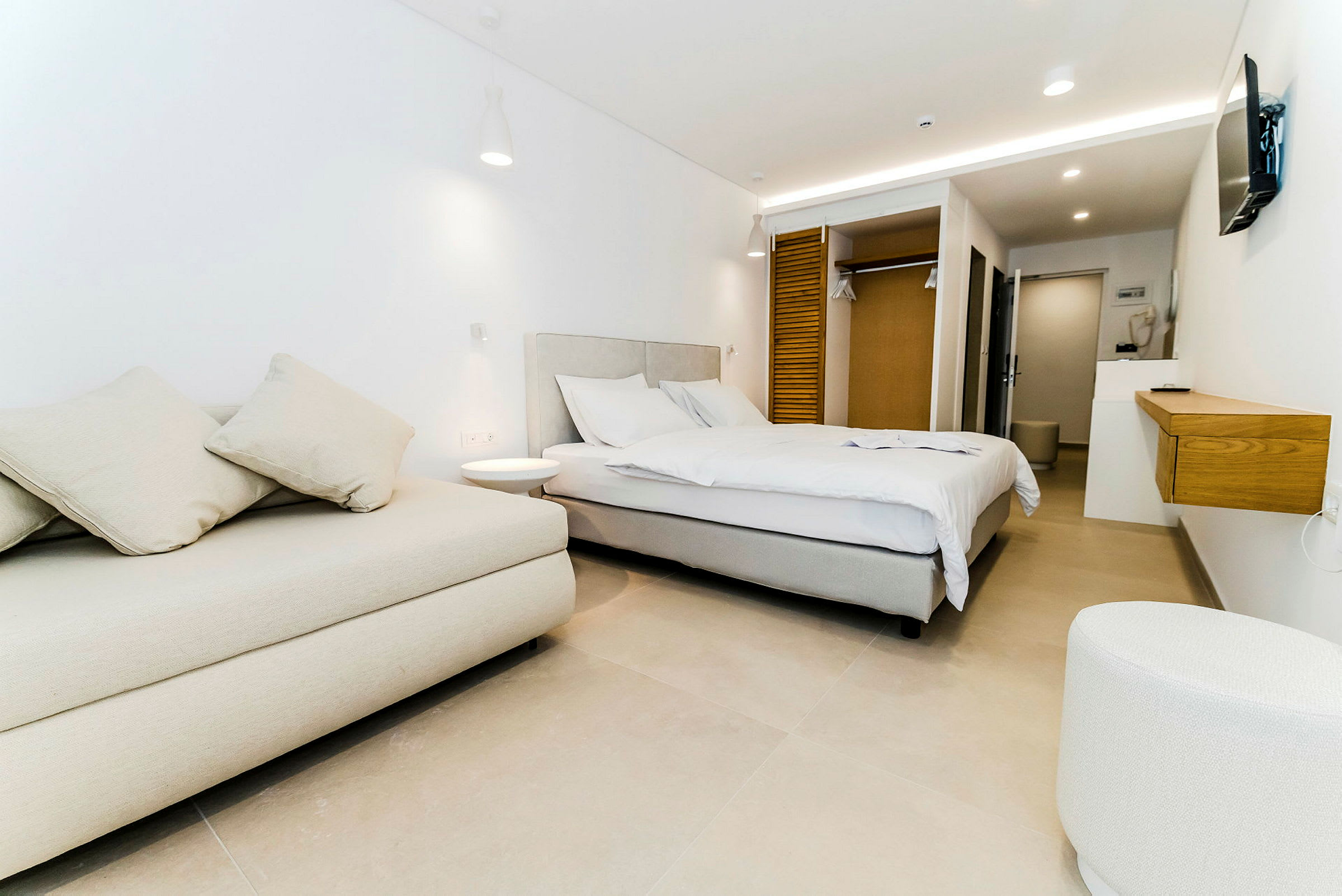 Et moderne hotellrom med en stor seng, en lys sofa, hvite vegger og en innebygd garderobe. (AI-generert tekst)