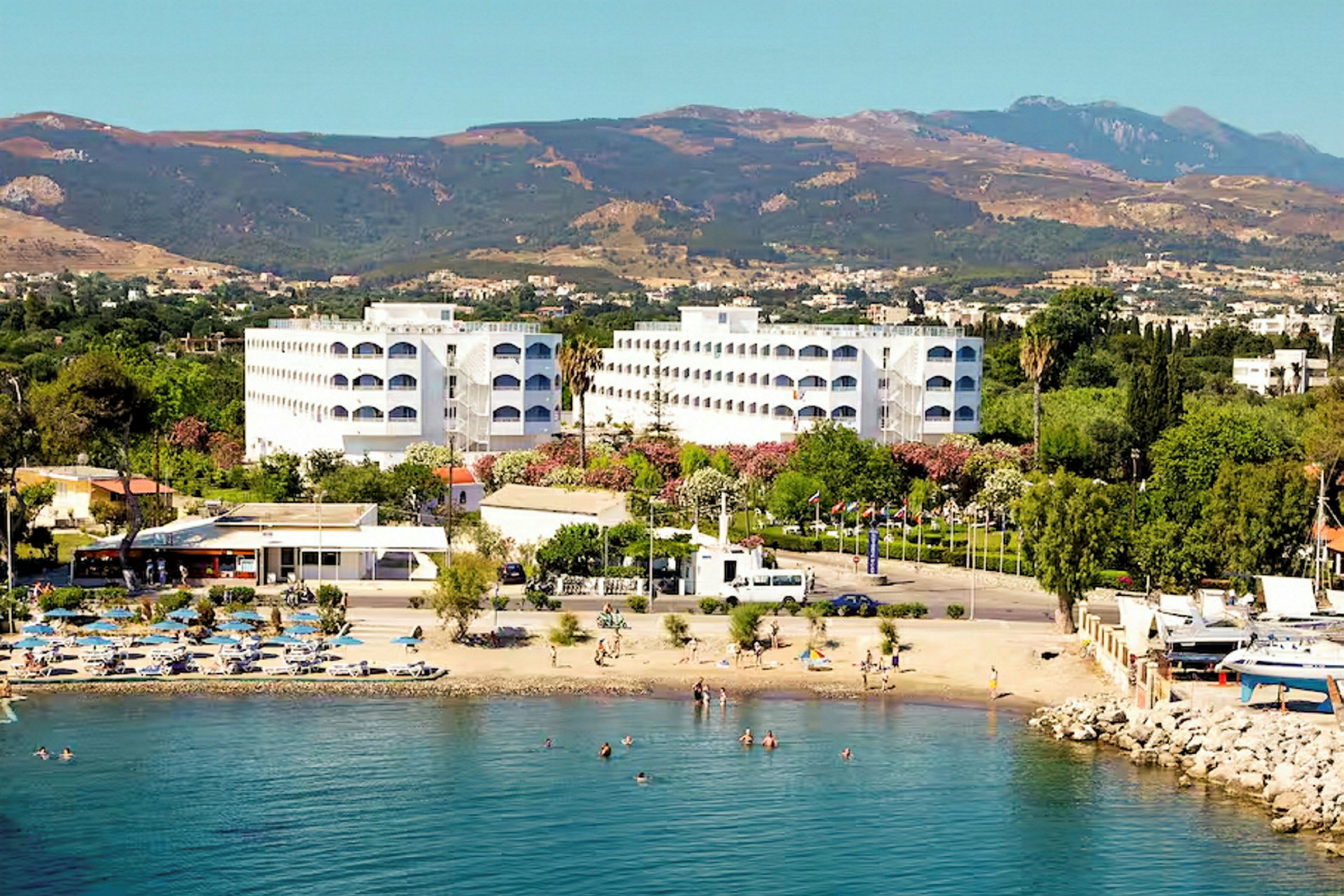 Hotellbygninger ved en strand med badegjester og fjell i bakgrunnen. (AI-generert tekst)