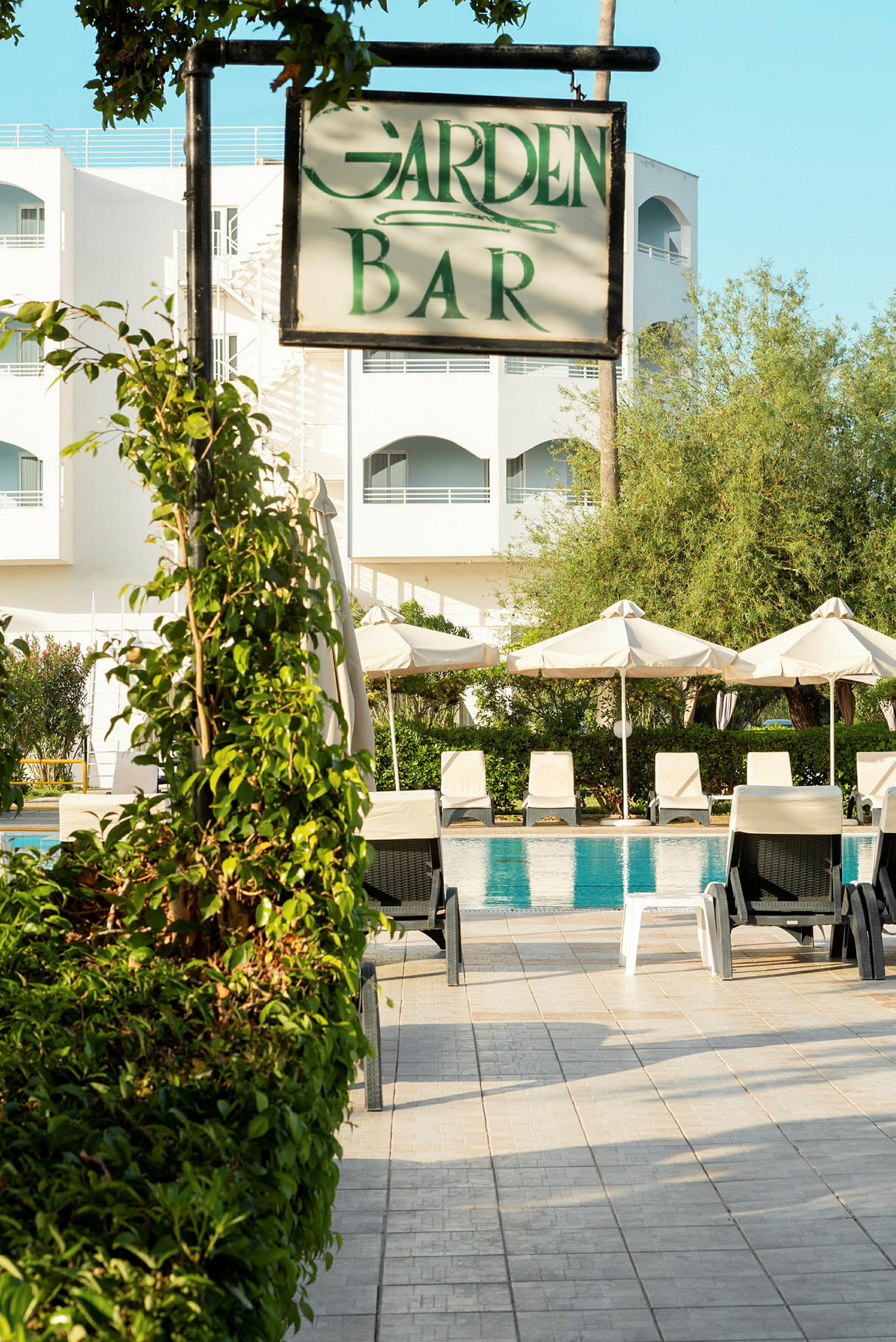 Skilt som viser 'Garden Bar' foran bassengområde og hotellbygning. (AI-generert tekst)