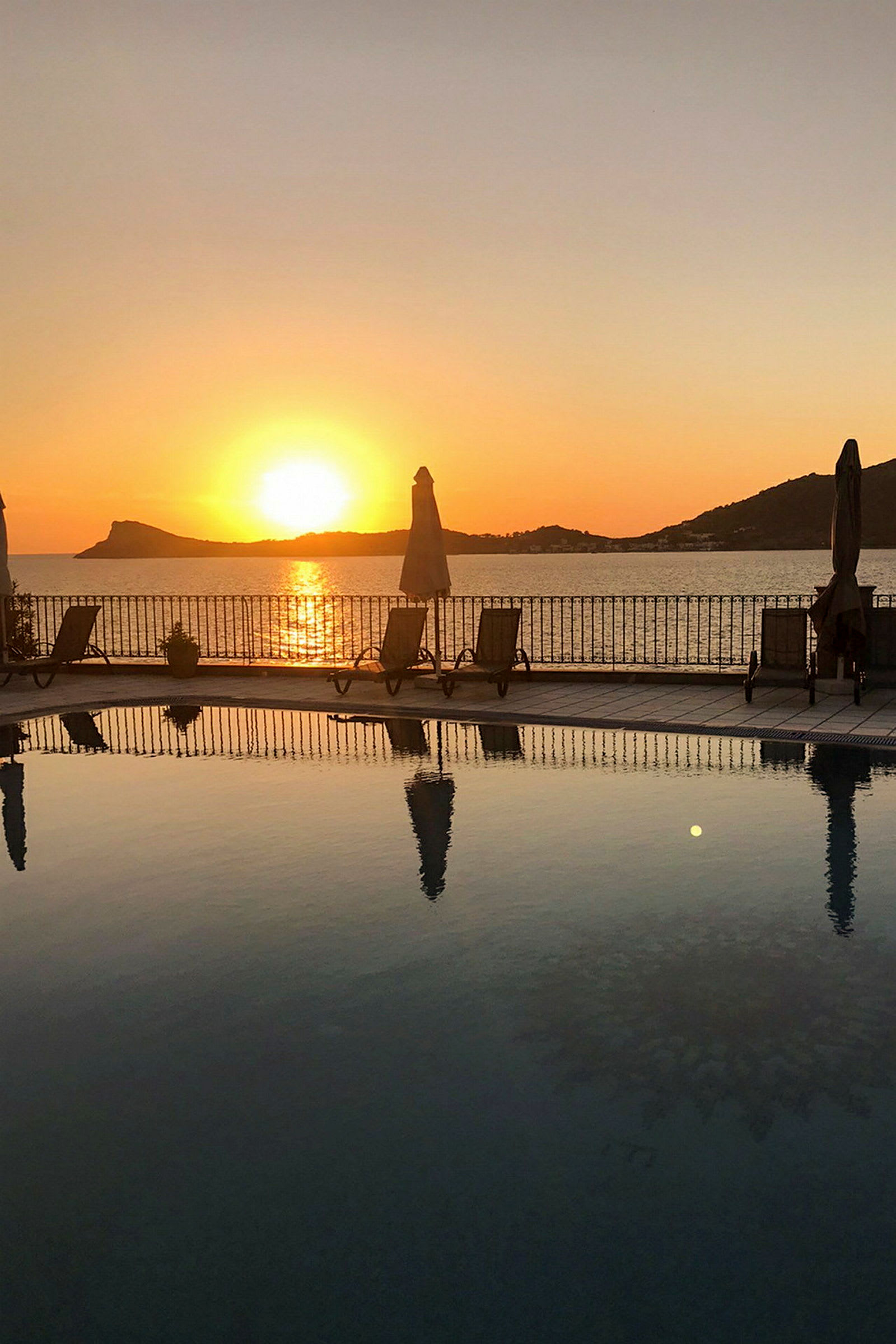 En solnedgang over havet med et basseng og solsenger i forgrunnen. (AI-generert tekst)