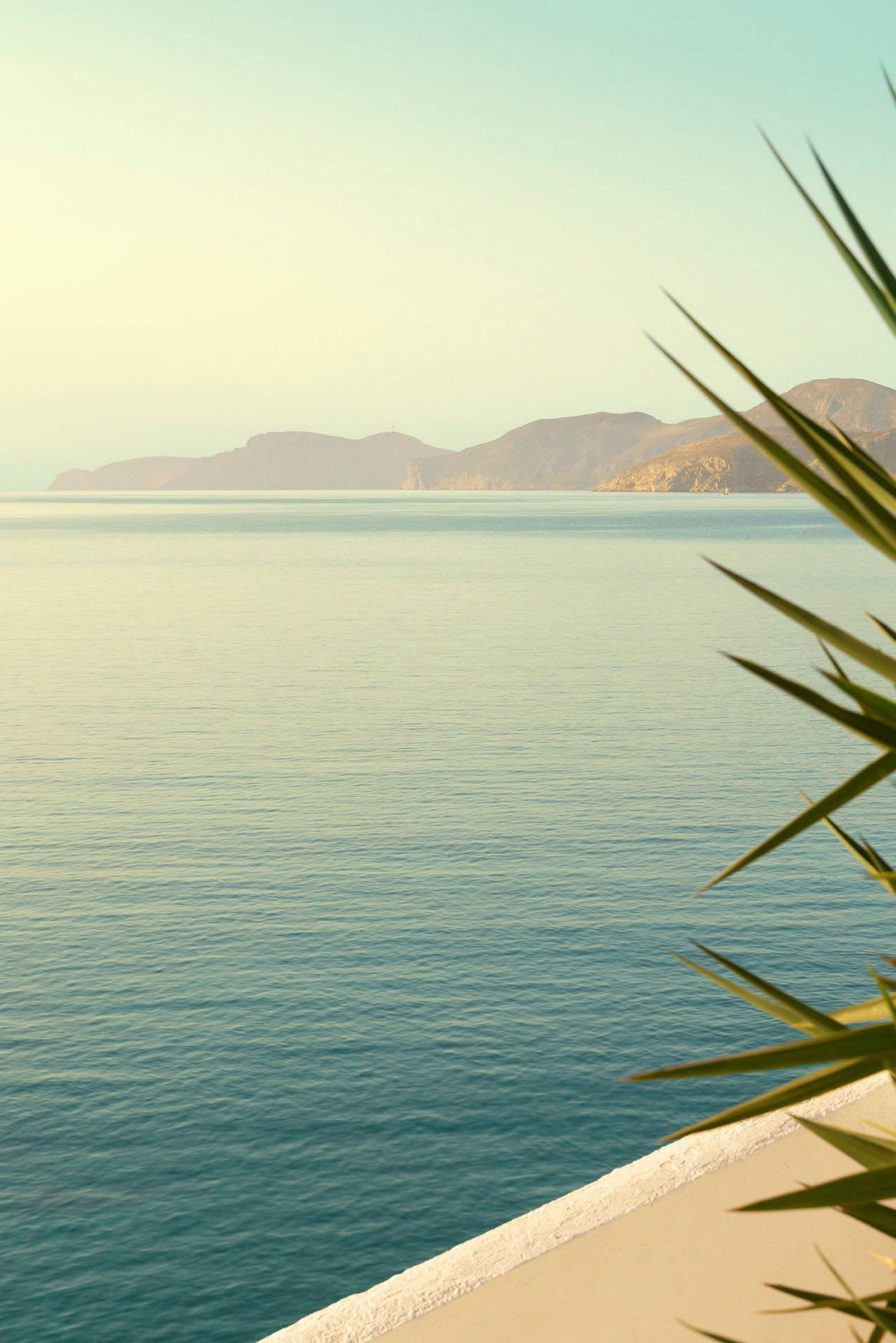 Bildet viser et blått hav med noen fjell synlige i bakgrunnen. Stranden er delvis synlig, og nær hjørnet er det grønne planter. (AI-generert tekst)