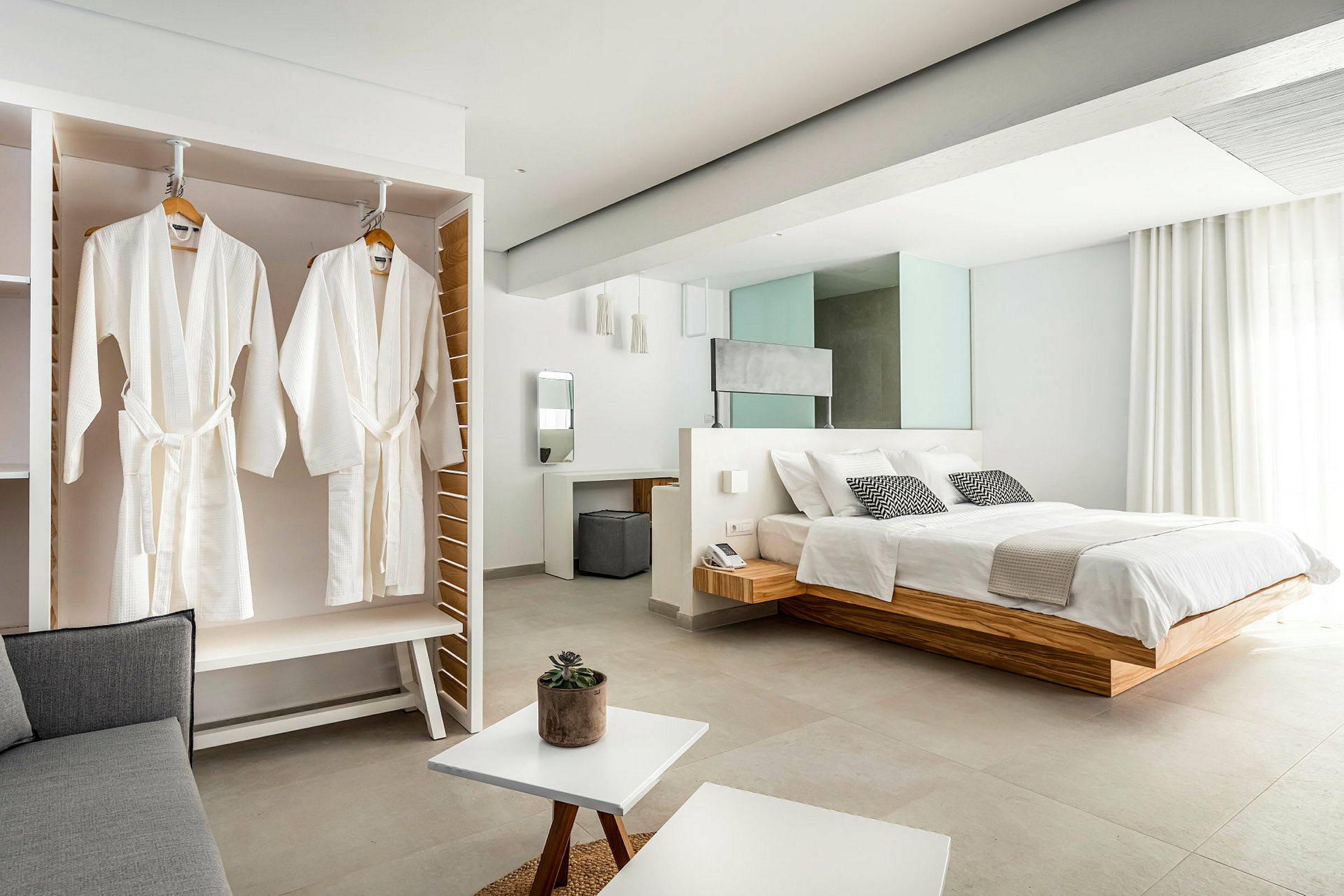 Bilde av et elegant hotellrom med en stor seng, hvite badekåper som henger i et skap og lyse moderne møbler. (AI-generert tekst)