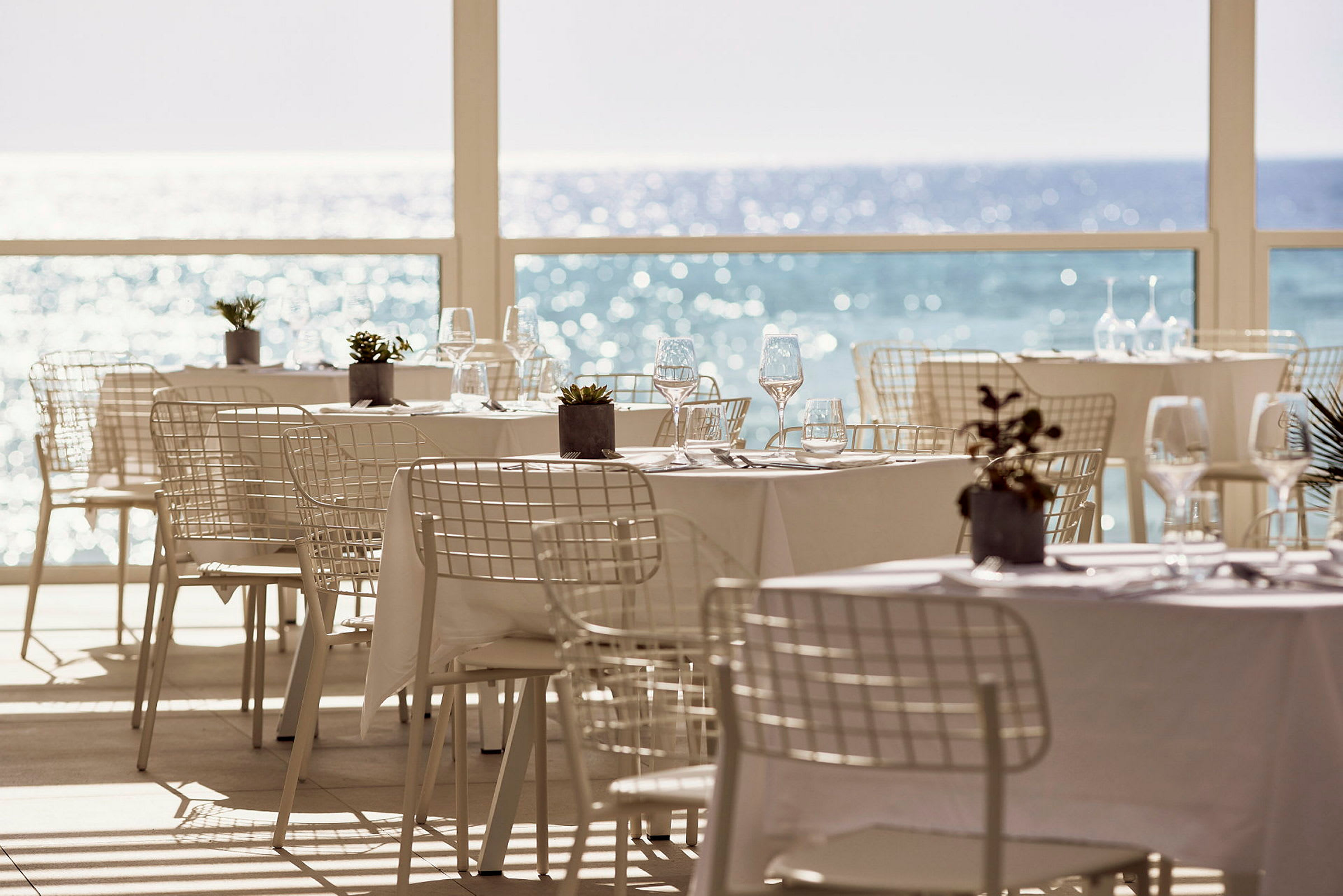 Restaurantbord med hvite duker og stoler nær et stort vindu med utsikt over havet. (AI-generert tekst)