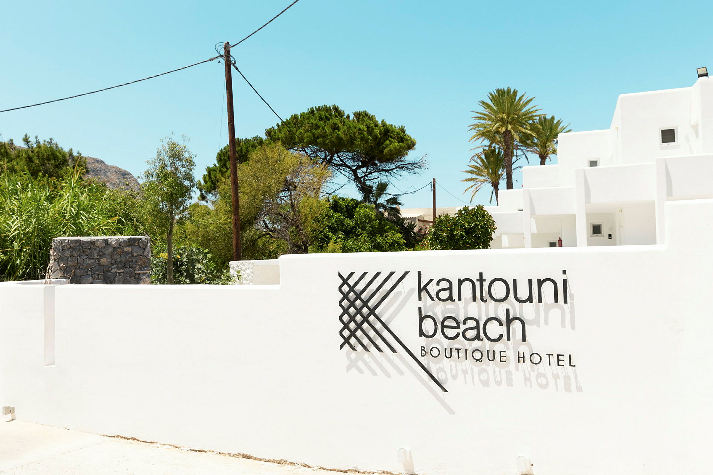 Bilde av en hvit bygning med teksten 'Kantouni Beach Boutique Hotel' på muren foran. Grøntområder og klar himmel i bakgrunnen. (AI-generert tekst)