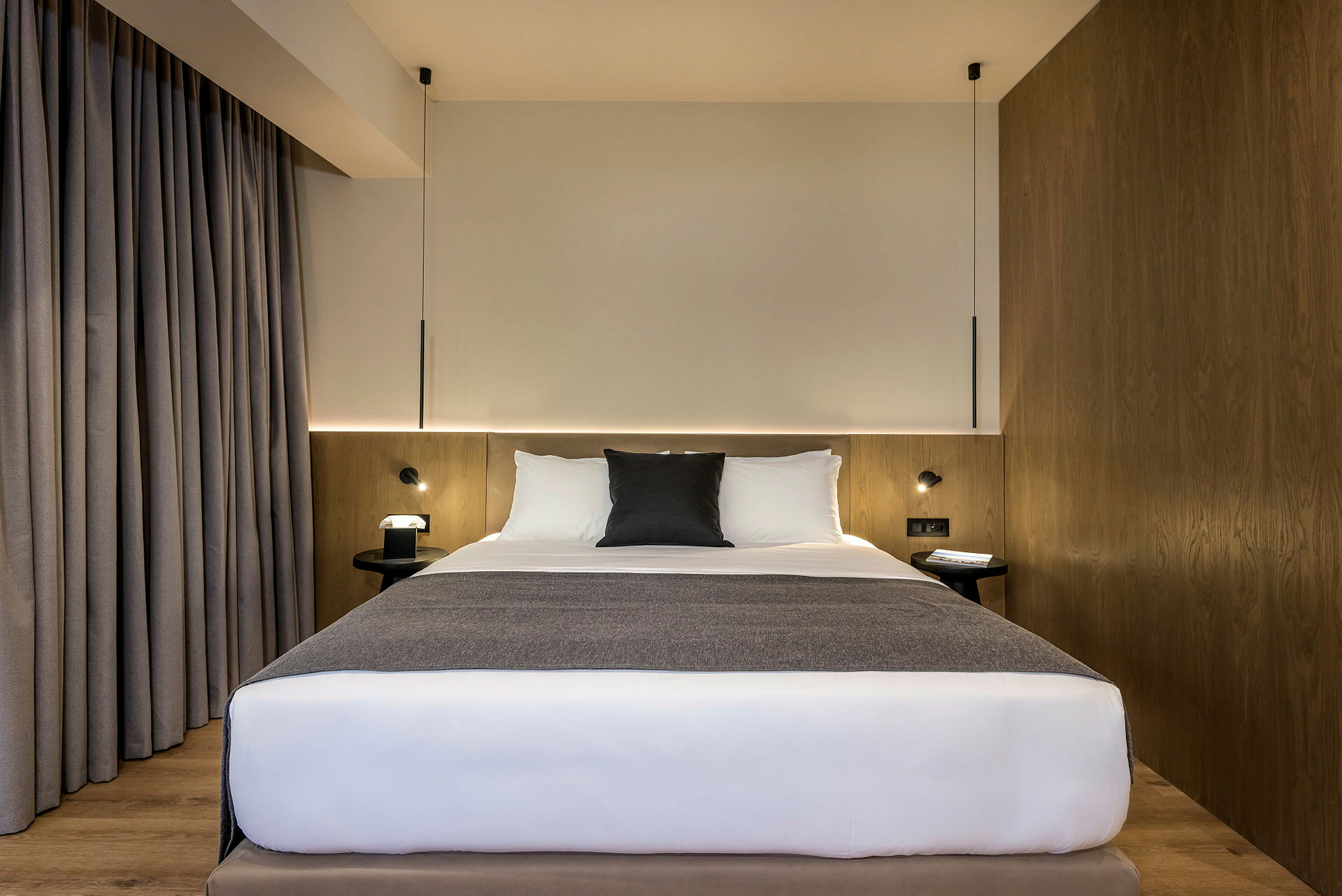 Et hotellrom med en seng med hvite laken og grå detaljer, trepanel og grå gardiner. (AI-generert tekst)