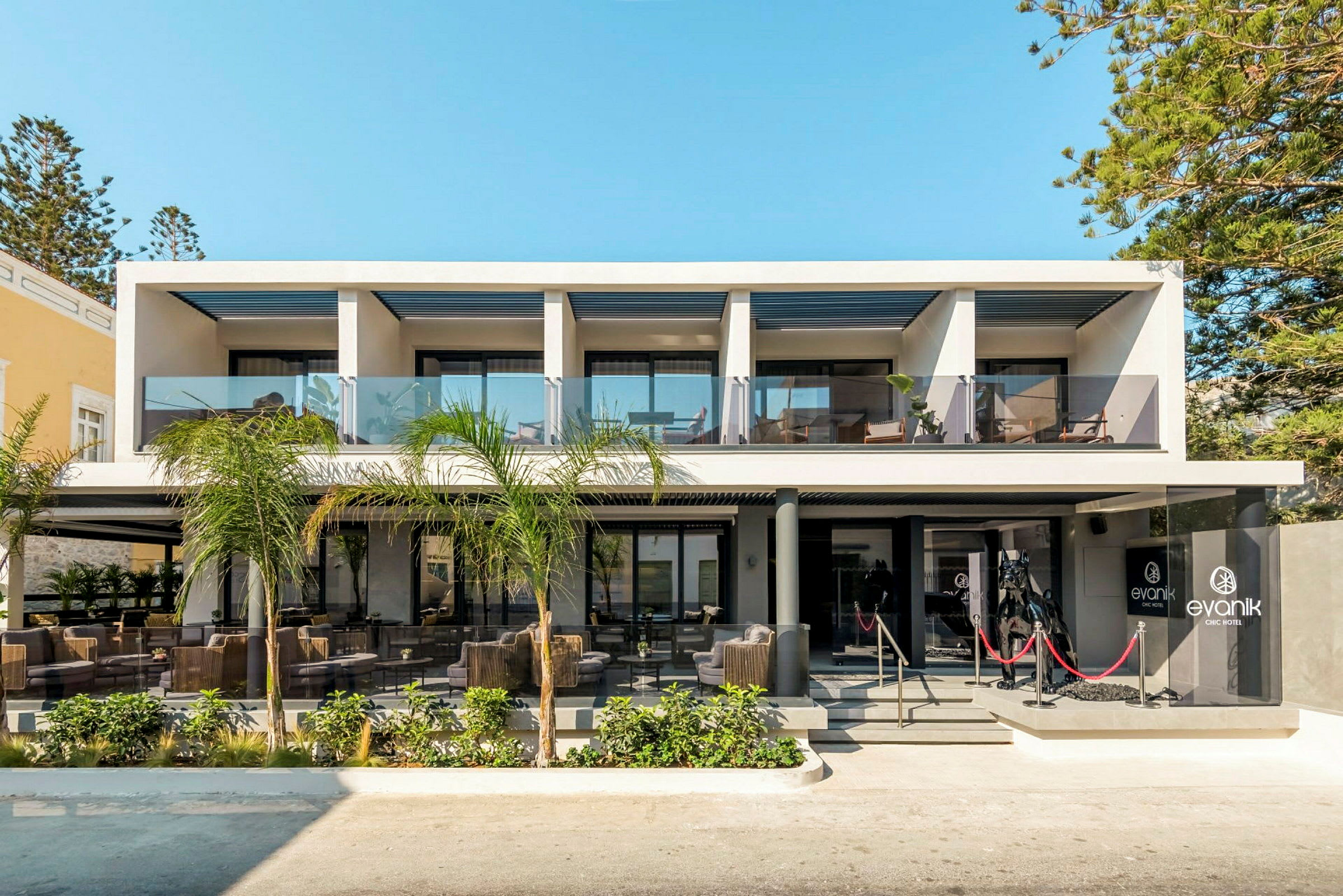 Moderne hotellbygning med glassrekkverk og utemøbler ved inngangen. (AI-generert tekst)