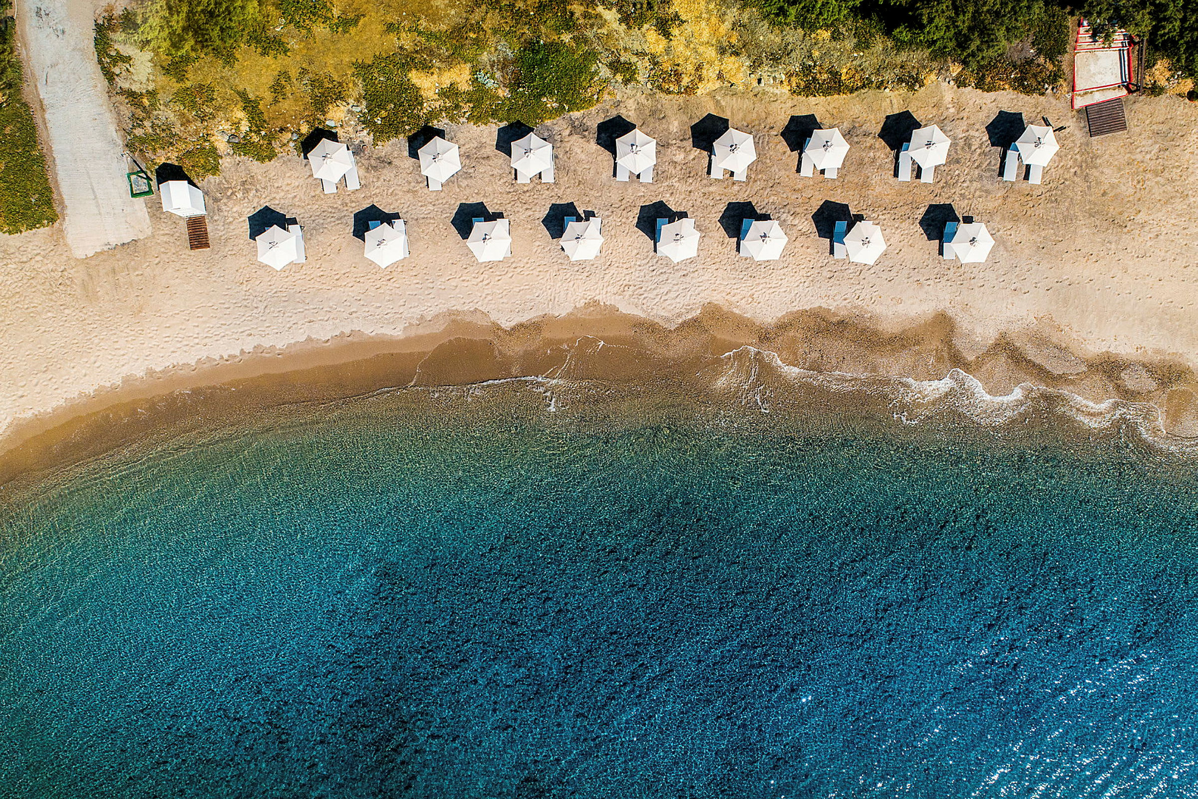 Luftfoto av en sandstrand med parasoller i rette rader og klart blått vann. (AI-generert tekst)
