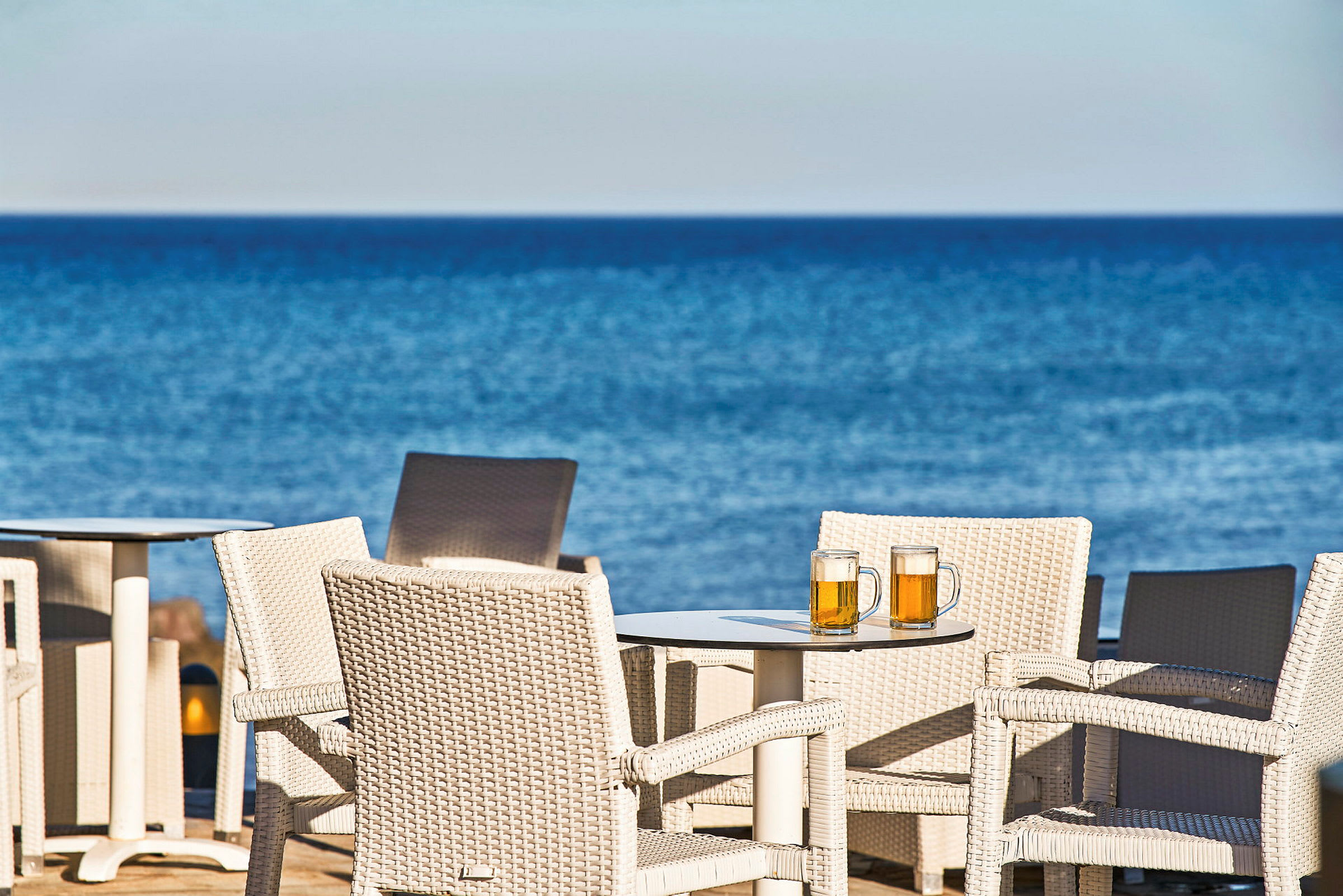 Kurvstoler ved et bord med to ølglass, med havet i bakgrunnen. (AI-generert tekst)