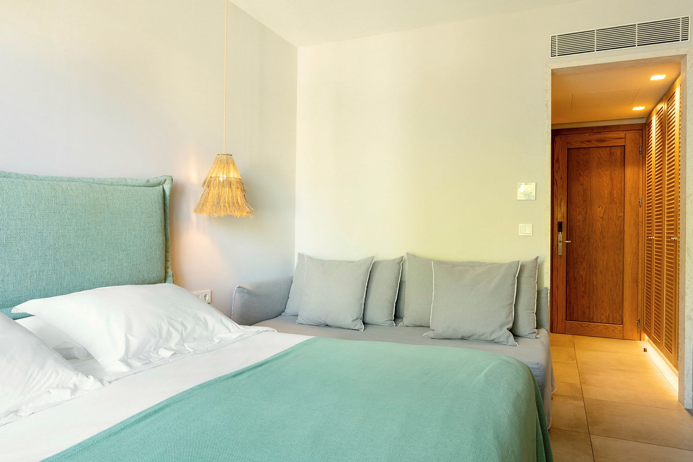 Et hotellrom med en seng med turkist sengetøy og hvite puter, samt en nærliggende sofa med grå puter. (AI-generert tekst)