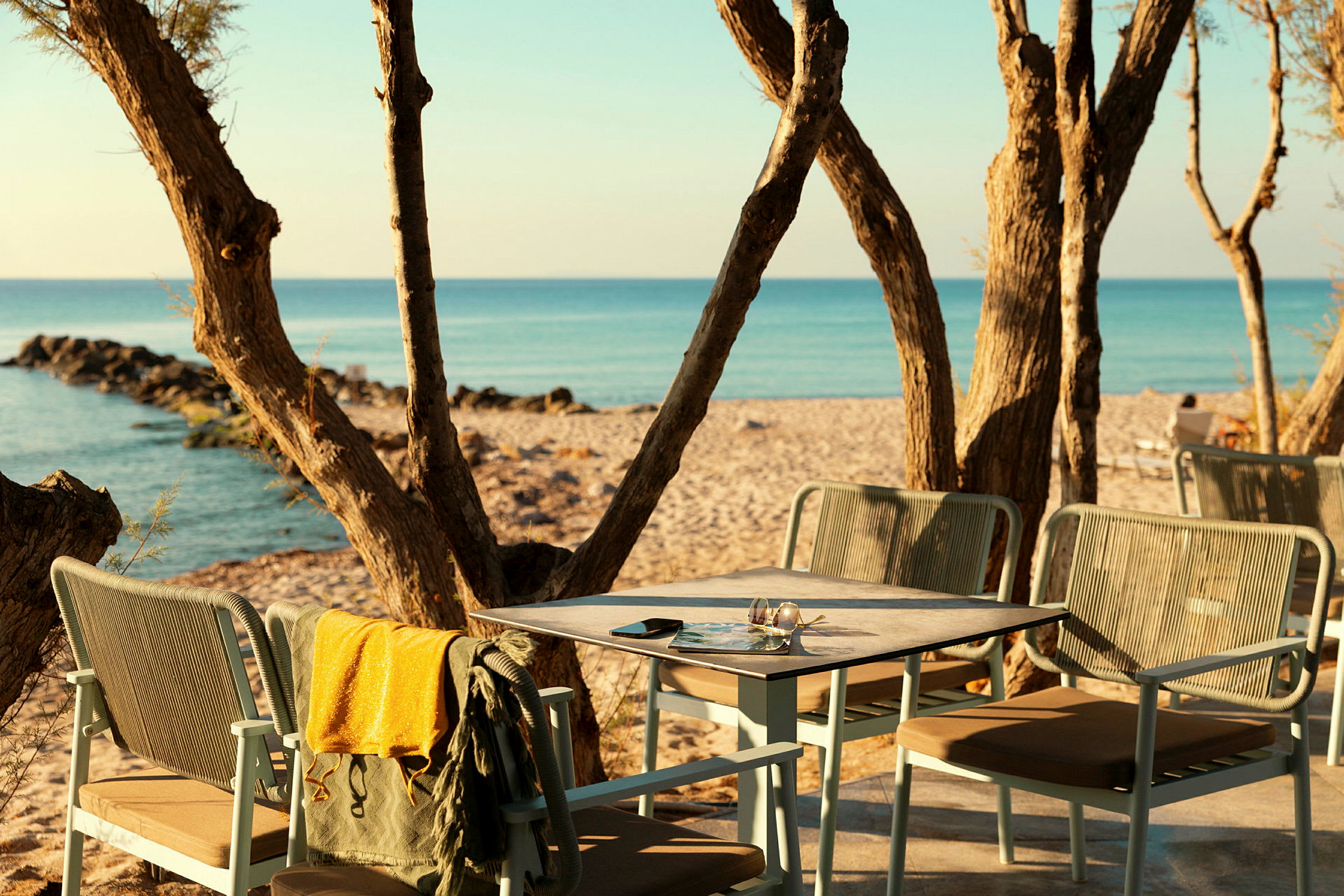 En strandscene med et bord og stoler på en terrasse blant trær, samt utsikt over havet og sandstranden. (AI-generert tekst)