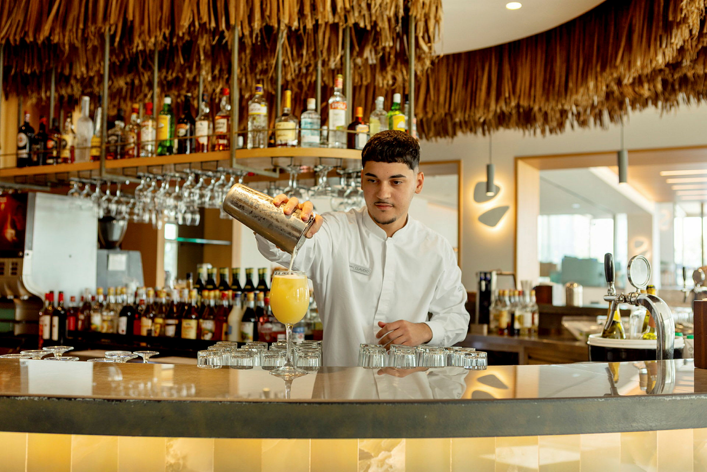 En bartender heller drikken i et glass foran en hylle med forskjellige brennevinsflasker og vinglass. (AI-generert tekst)