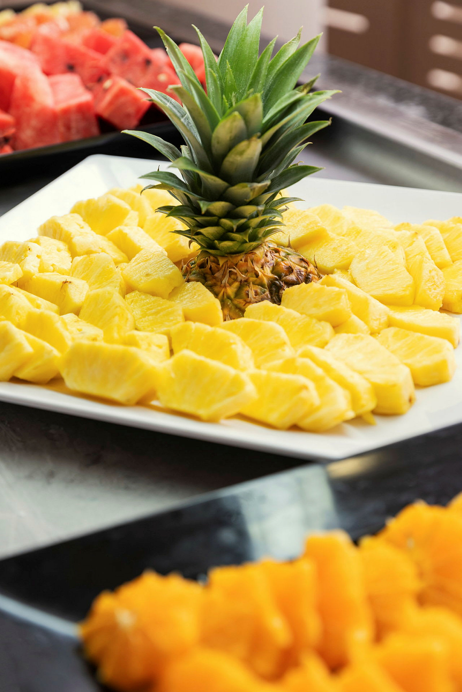 Et nærbilde av en tallerken med skiver av fersk ananas og dens grønne krone, med andre frukter i bakgrunnen. (AI-generert tekst)