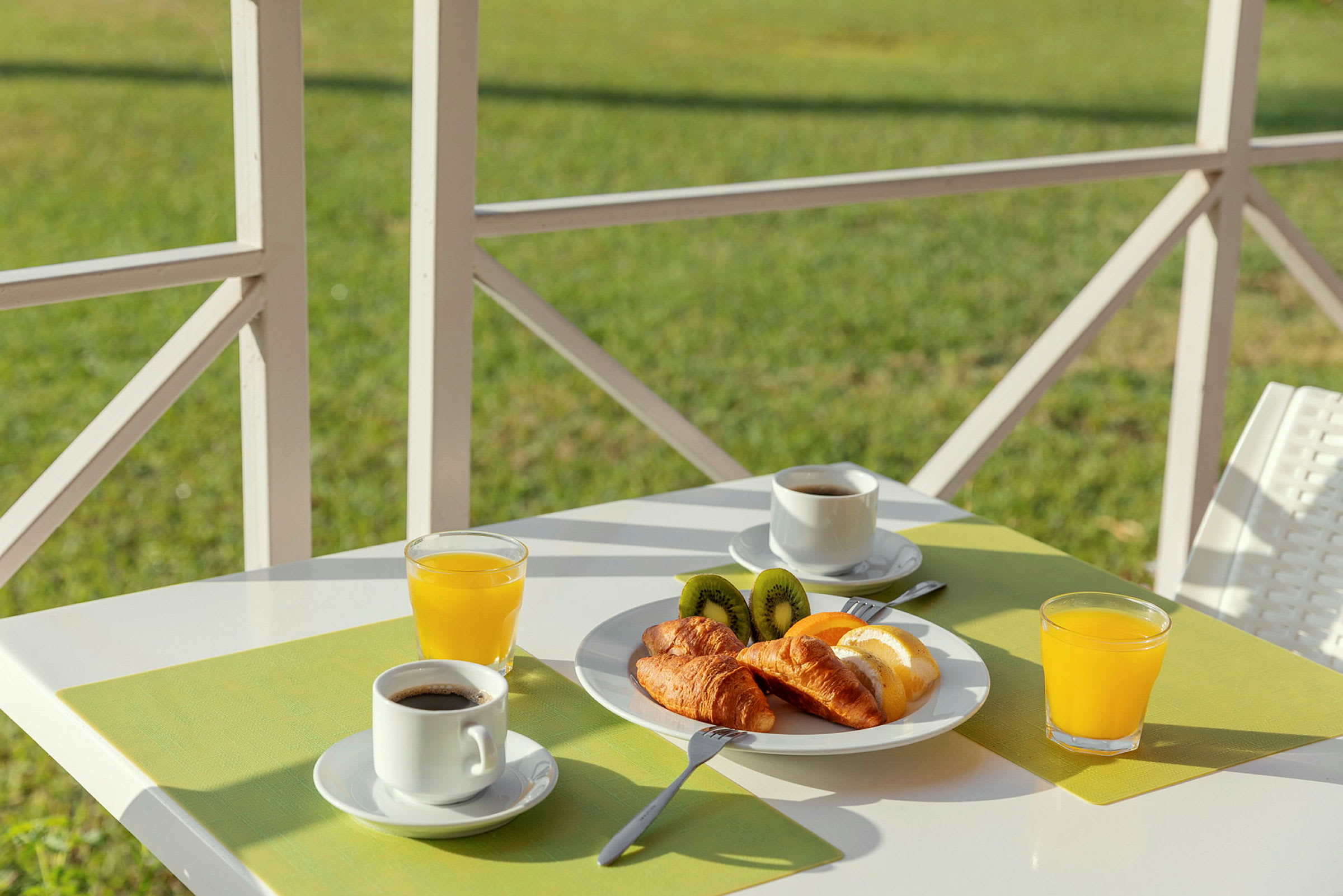 Frokost med kaffe, juice, croissanter og kiwi på et hvitt bord med grønn bordbrikke. (AI-generert tekst)