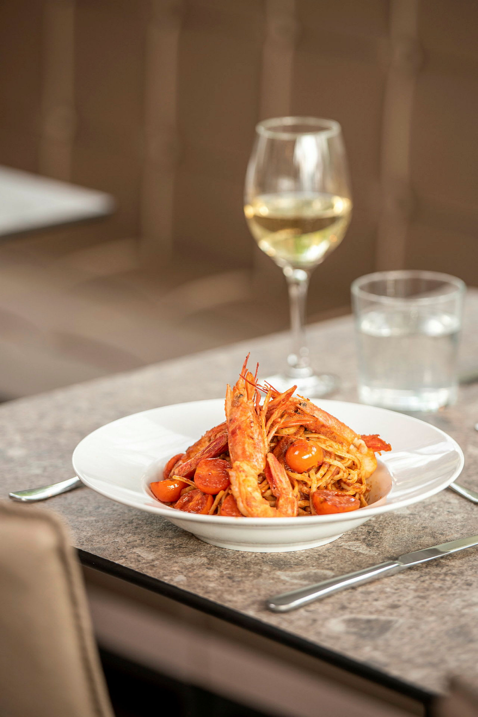 En tallerken med pasta, reker og tomater står på et bord. I bakgrunnen vises et glass hvitvin. (AI-generert tekst)
