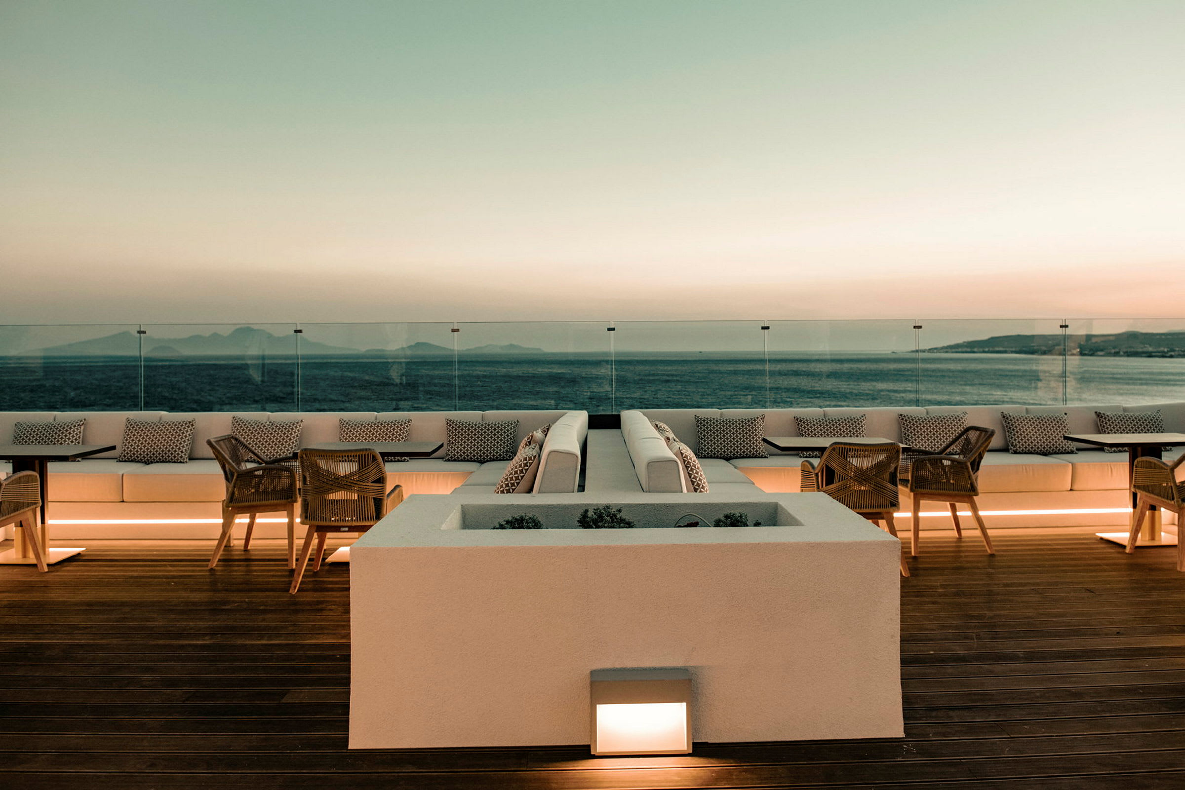 En terrasse med sitteplasser og bord med utsikt over havet ved solnedgang. (AI-generert tekst)