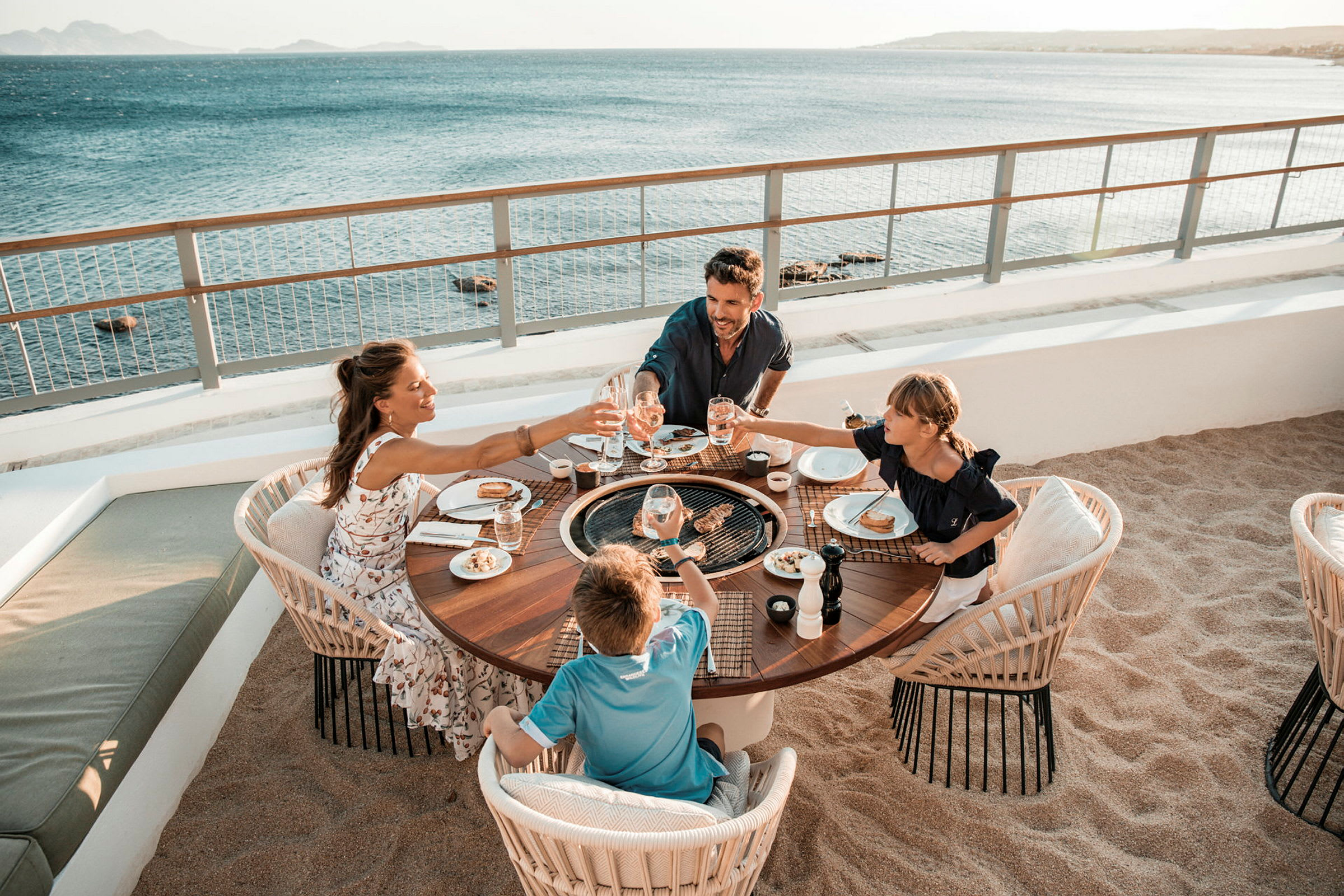 En familie sitter ved et rundt bord utendørs ved stranden og nyter et måltid med utsikt over havet. (AI-generert tekst)