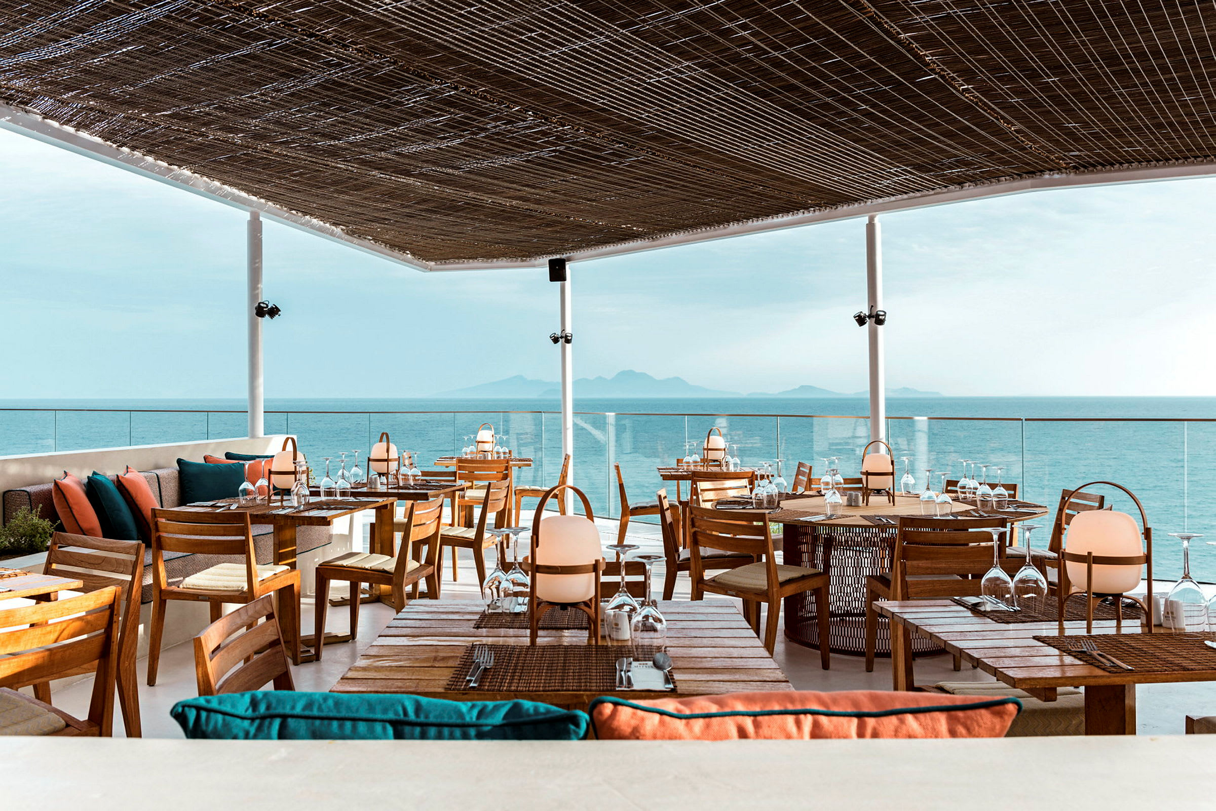 En restaurant med takterrasse med utsikt over havet. Møblene er laget av tre med fargerike puter, og taket består av et flettet materiale som slipper inn lys. (AI-generert tekst)
