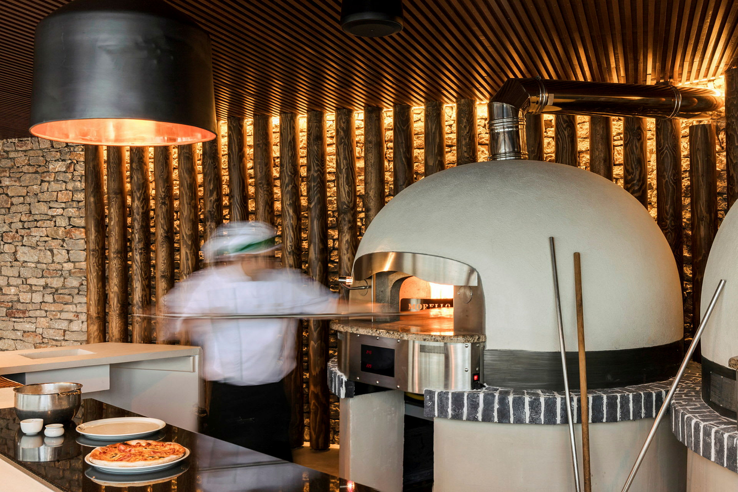 En pizzabaker foran en vedfyrt ovn med et moderne kjøkken i bakgrunnen. (AI-generert tekst)