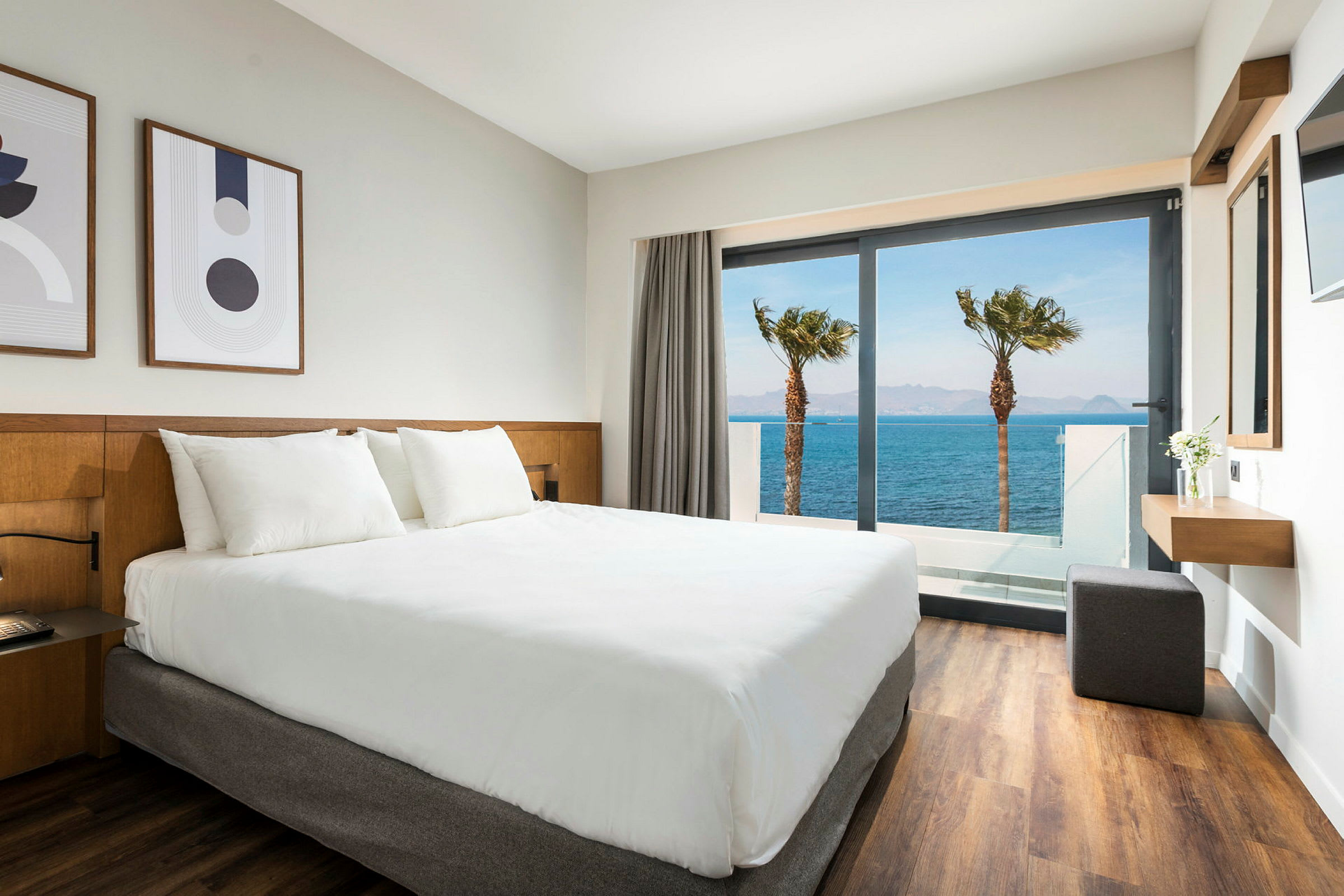 Hotellrom med hvit seng og utsikt over havet gjennom en glassdør. (AI-generert tekst)