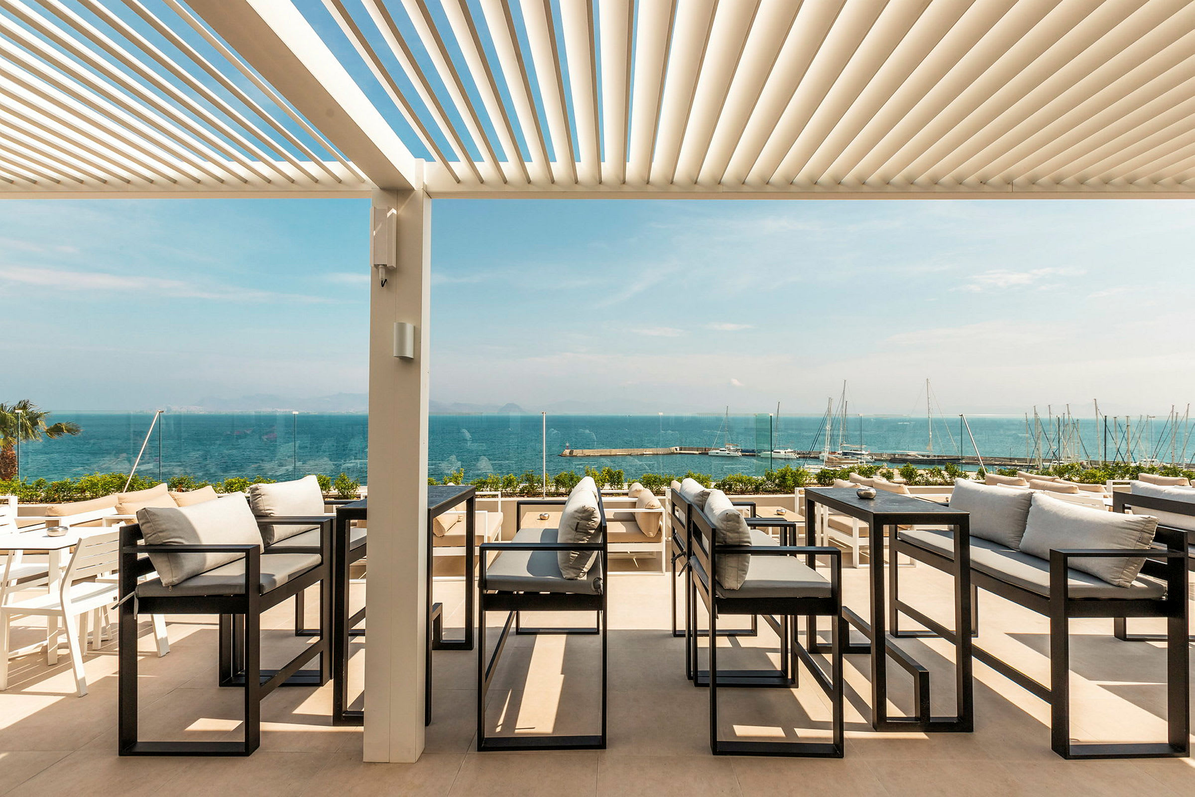 Utendørs servering med utsikt over havet, med sofaer og bord under en pergola. (AI-generert tekst)