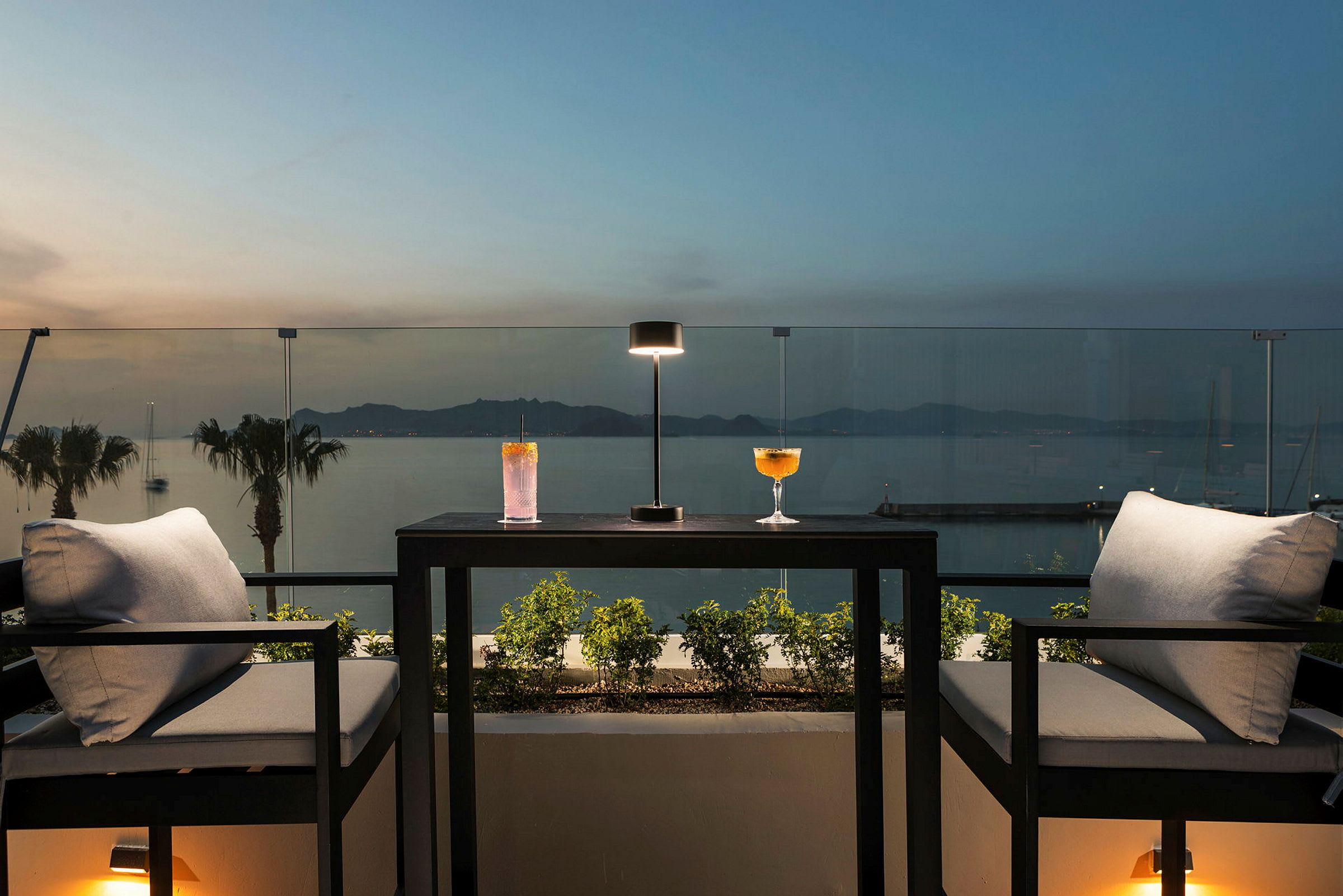 To lenestoler med et bord mellom, med to drinker og en lampe på bordet, foran et glassrekkverk med utsikt over havet i skumringen. (AI-generert tekst)