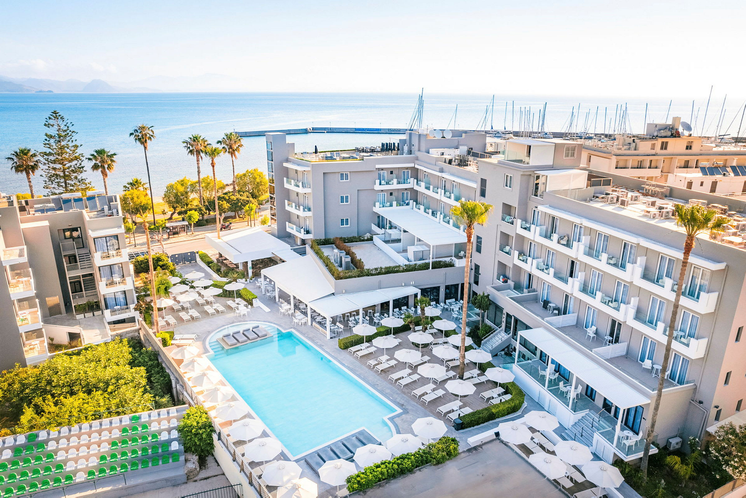 Et hotellområde med basseng, parasoller og solsenger omgitt av bygninger. Hotellet ligger nær sjøen med en strand og en småbåthavn i bakgrunnen. (AI-generert tekst)