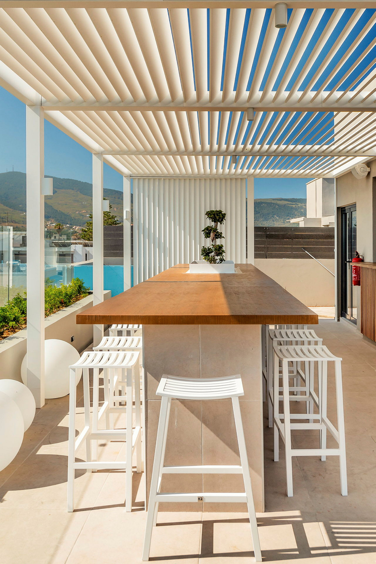 Uteservering med et stort trebord omgitt av hvite barkrakker og en pergola for skygge. Fjelllandskapet kan skimtes i bakgrunnen. (AI-generert tekst)