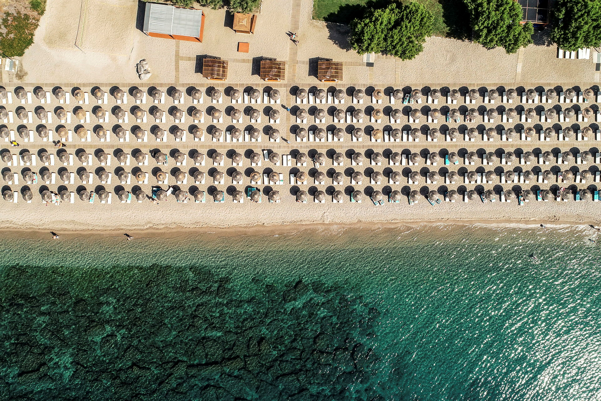 Flyfoto av en strand med parasoller og solsenger i symmetriske rekker ved sjøkanten. (AI-generert tekst)