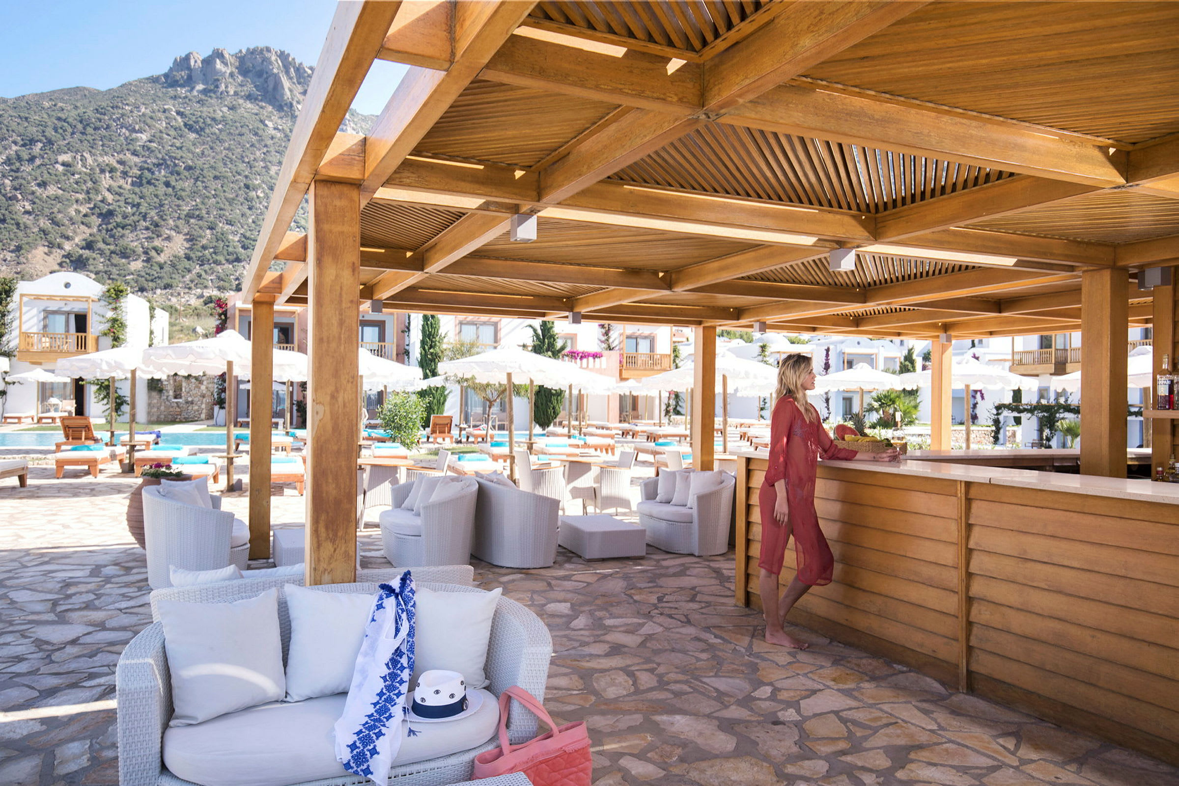 Bassengområde med bar og sitteplasser under en trepergola. Fjell i bakgrunnen. (AI-generert tekst)