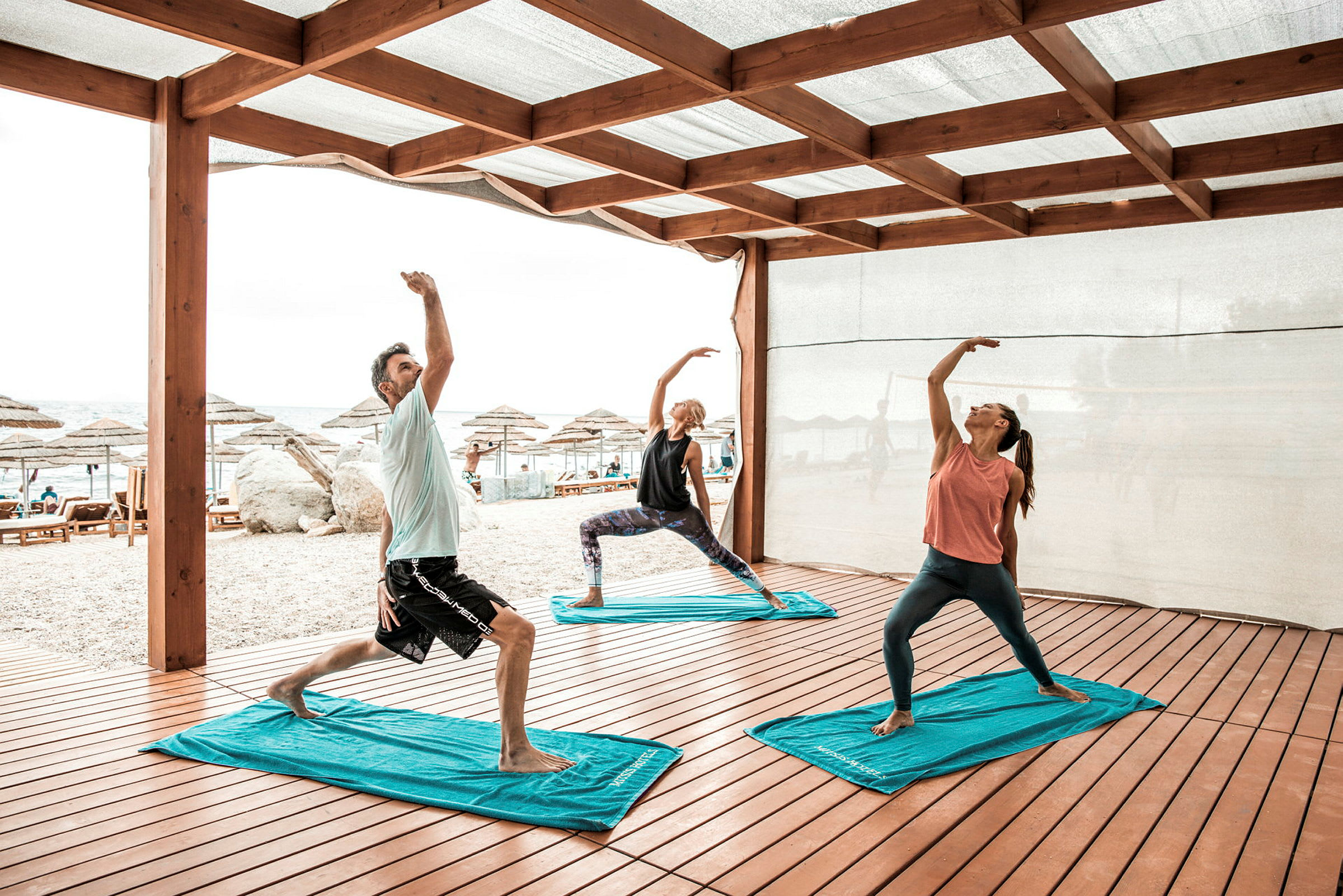 Tre personer utfører yogaøvelser under en trekonstruksjon nær en strand. (AI-generert tekst)